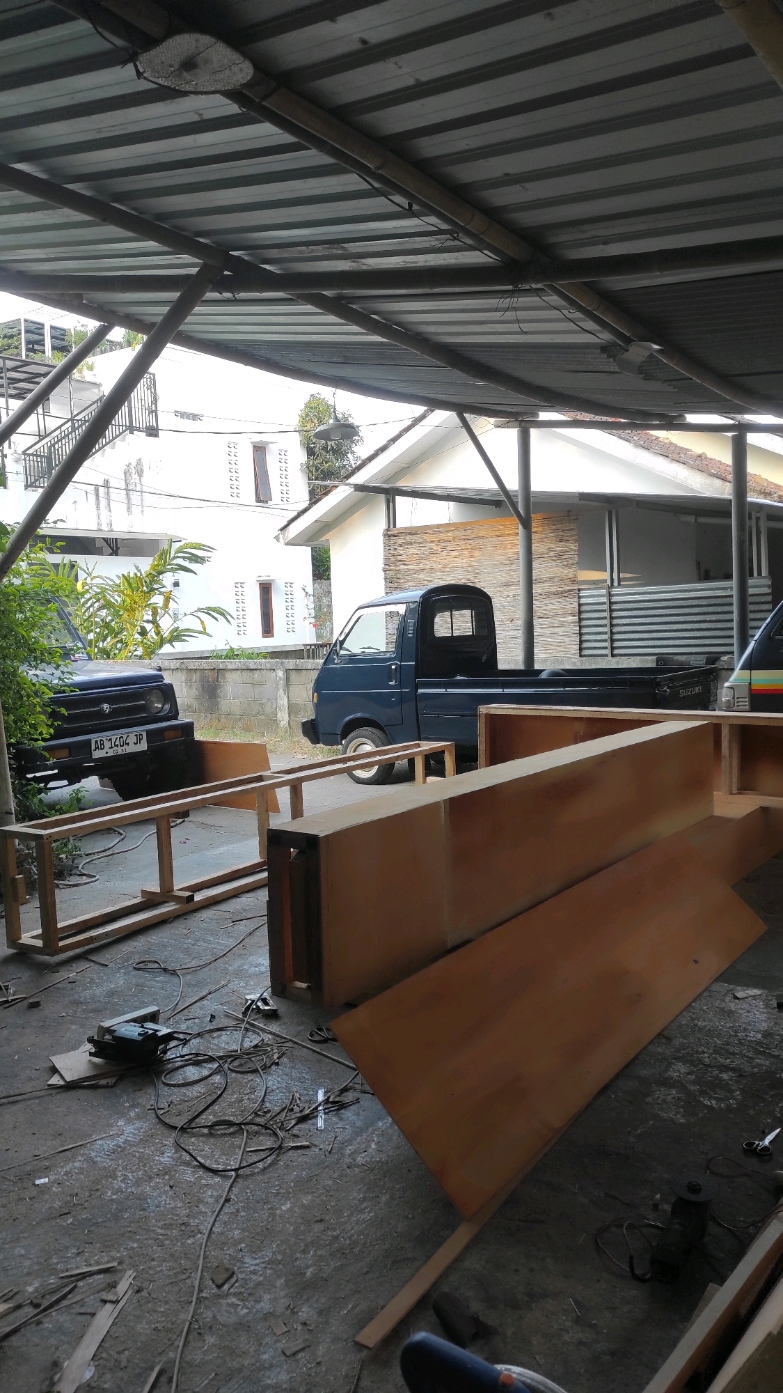 The image depicts an outdoor workshop area, likely a backyard or covered patio, in Guwosari, Indonesia. The scene is partially covered by a corrugated metal roof supported by bamboo and metal framing, suggesting a basic structure for shelter. In the foreground, unfinished wooden furniture pieces, possibly shelving units, are laid out on the concrete ground. Various tools and materials are scattered around, including an electric drill with its cord, scissors, and scraps of wood, indicating ongoing construction or assembly. Two blue vehicles are parked side-by-side in the background: a dark blue Suzuki Jimny 4x4 on the left and a dark blue utility pickup truck on the right. Behind the vehicles, a white house with patterned windows and a second structure with a corrugated metal facade are visible, along with some greenery. The overall impression is of a functional, albeit somewhat cluttered, workspace engaged in furniture construction.