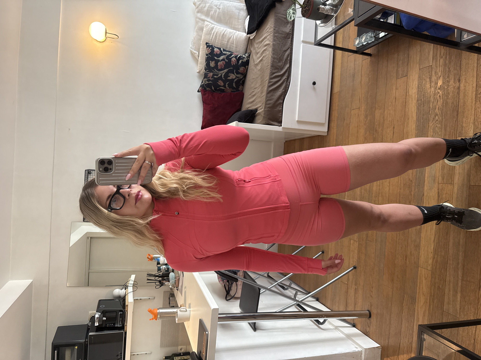 A woman with blonde hair and glasses stands in a room, taking a mirror selfie. She is wearing a coral-colored athletic outfit consisting of a zip-up jacket and shorts. Her phone, in a silver case, is held up to take the picture.
The background shows a room with light-colored walls and wooden flooring. To the left, a shelf holds various kitchen appliances, including a black microwave and another black appliance. A white table is also visible, with a mug and a metal dispenser on it. In the upper left corner, a small, circular light fixture is mounted on the wall.
Further back, a bed is partially visible, covered with pillows and blankets. The overall setting appears to be an indoor space, possibly a studio apartment or a living area. The lighting suggests it could be daytime, though the exact time is not discernible. No specific weather conditions are indicated. There is no visible text in the image. The scene is casual and appears to be a personal moment captured by the woman.