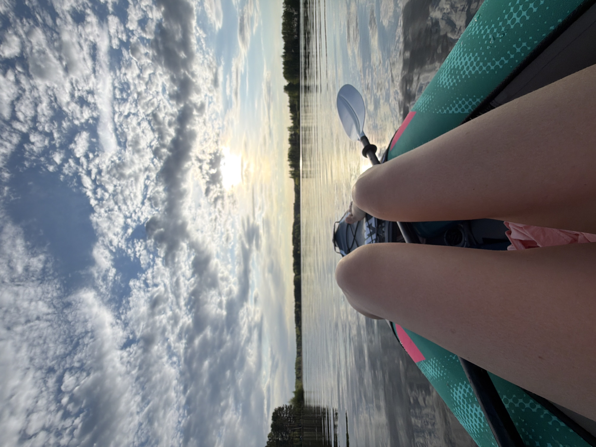 A first-person perspective shot from a boat or kayak, presumably on the Kama River near Chaykovsky, Russia. The focus is on the kayaker's thighs and the kayak itself, with a paddle resting nearby. The background shows a view of the river, the opposite bank with trees, a cloudy sky, and the sun shining through.