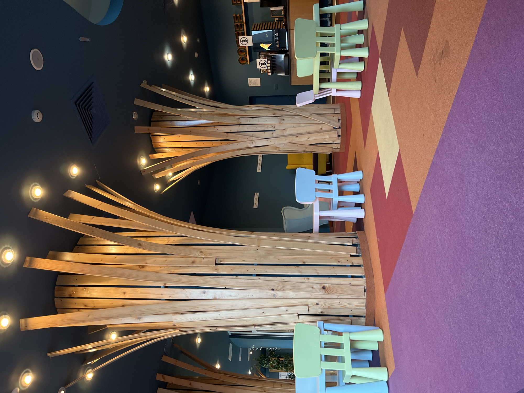 The image shows a brightly colored children's play area with small tables and chairs. The area is decorated with tall, wooden columns that resemble trees, and the floor is covered with a patterned carpet in shades of purple, orange, red, and yellow. In the background, there is a coffee counter with a coffee machine and a sign that reads "COFFEE". The location is identified as being in the city of Nesebar, Bulgaria. The scene is well-lit with overhead lighting, and the atmosphere appears to be welcoming and child-friendly. There are no people visible in the image.