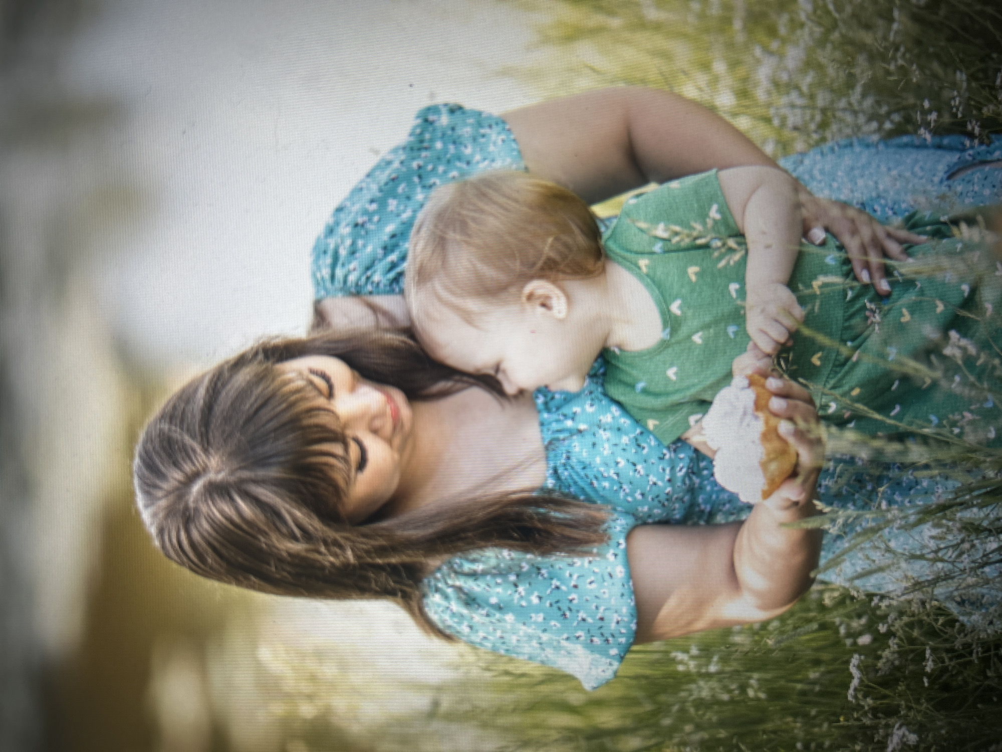 The image shows a woman holding a baby in an outdoor setting. The woman is wearing a blue floral dress and has long brown hair. The baby is wearing a green outfit with a pattern of small white shapes. The woman is looking down at the baby with a smile, and the baby is looking towards the woman. The baby is holding a small pastry in their hands. The background is blurred and appears to be a grassy area with some water in the distance. The lighting suggests it is daytime. The specific location in the city of Kiselyovsk, Russia, cannot be determined from the visual information provided in the image.