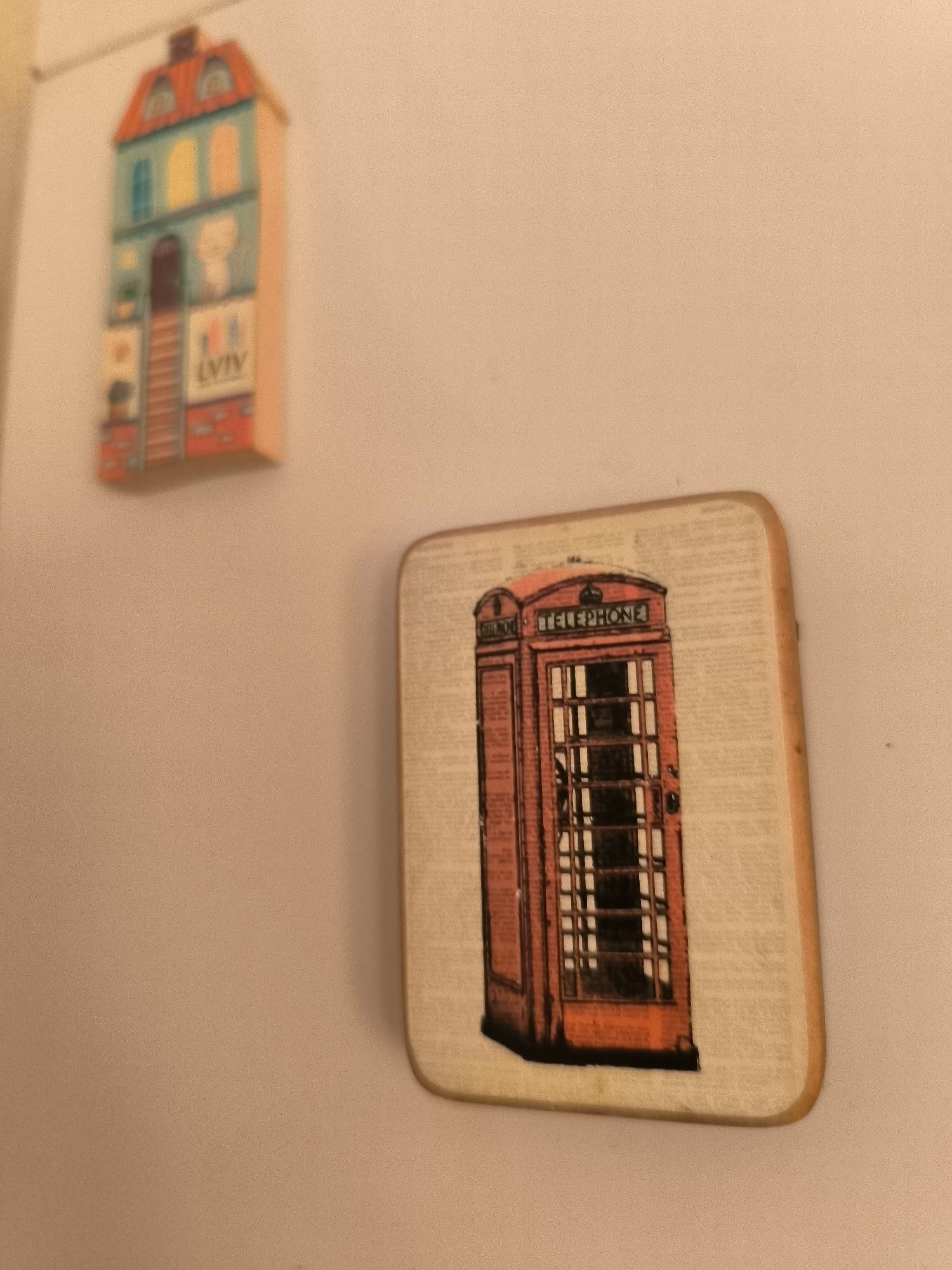 The image shows two magnets on a white surface, likely a refrigerator. The magnet on the left depicts a stylized building with a red roof and colorful windows, featuring the text "LVIV". The magnet on the right is square-shaped and features an illustration of a red British telephone box with the word "TELEPHONE" above it. The background is a neutral, light-colored surface. There is no indication of the city of Horishni Plavni, Ukraine, in the image.