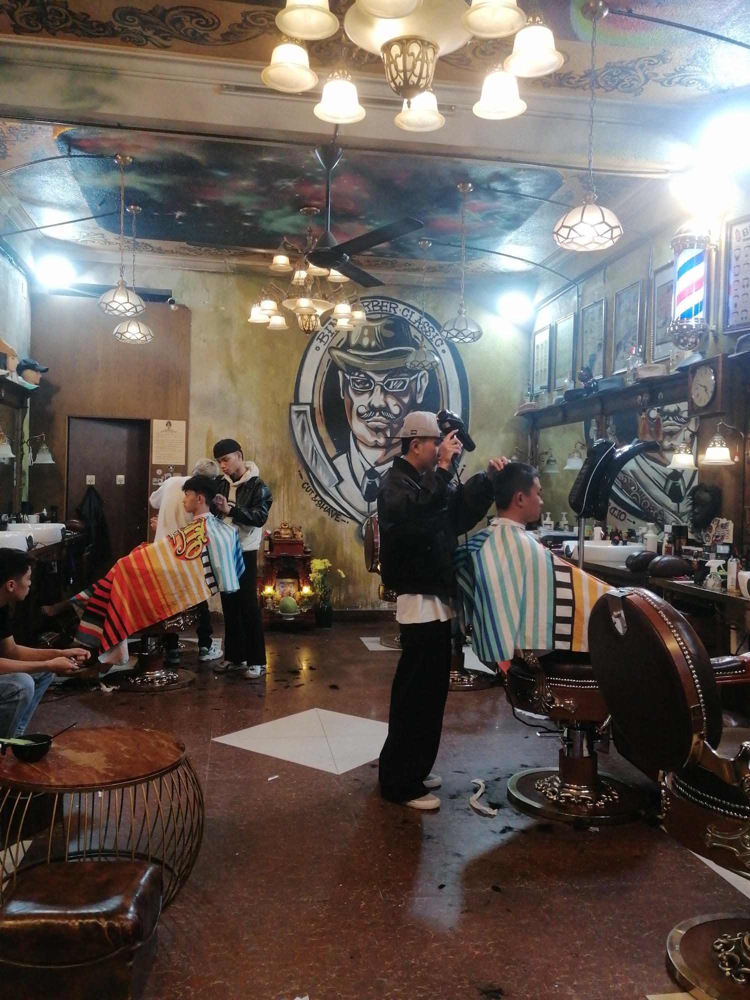 Inside a barber shop in Da Lat, Vietnam, are at least three barbers providing services to patrons.
One barber in a black jacket and backwards grey baseball cap is taking a picture of a customer with an electronic device.
Another barber is cutting hair for a customer.
A third person is sitting in a barber's chair, though it is not clear if they are a barber, a customer, or neither.
The shop contains ornate lighting fixtures, barber chairs, mirrors, a wall mural depicting a man wearing a fedora, and decorative items on shelves.
Hair clippings are scattered on the floor, indicating recent activity.