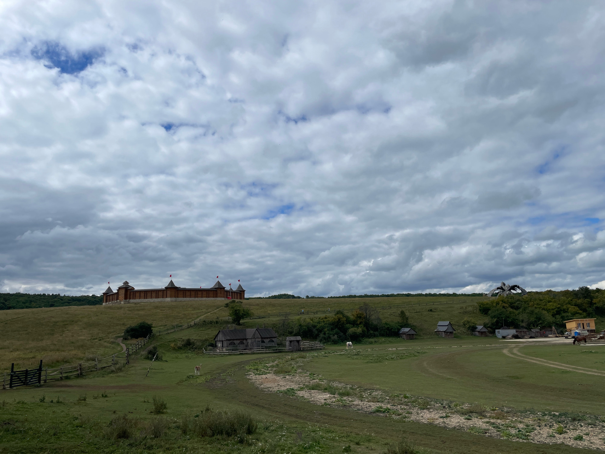 The image depicts a wide, grassy landscape under a cloudy sky. On a hill in the distance, a large, wooden fortress with red flags on its towers is visible. In the mid-ground, there are several small wooden buildings, a white horse grazing in a field, and a fence that winds through the grass. A dirt path leads up to the fortress. In the far right of the image, a brown horse is being ridden on a dirt track. The overall scene suggests a historical or reenactment site.