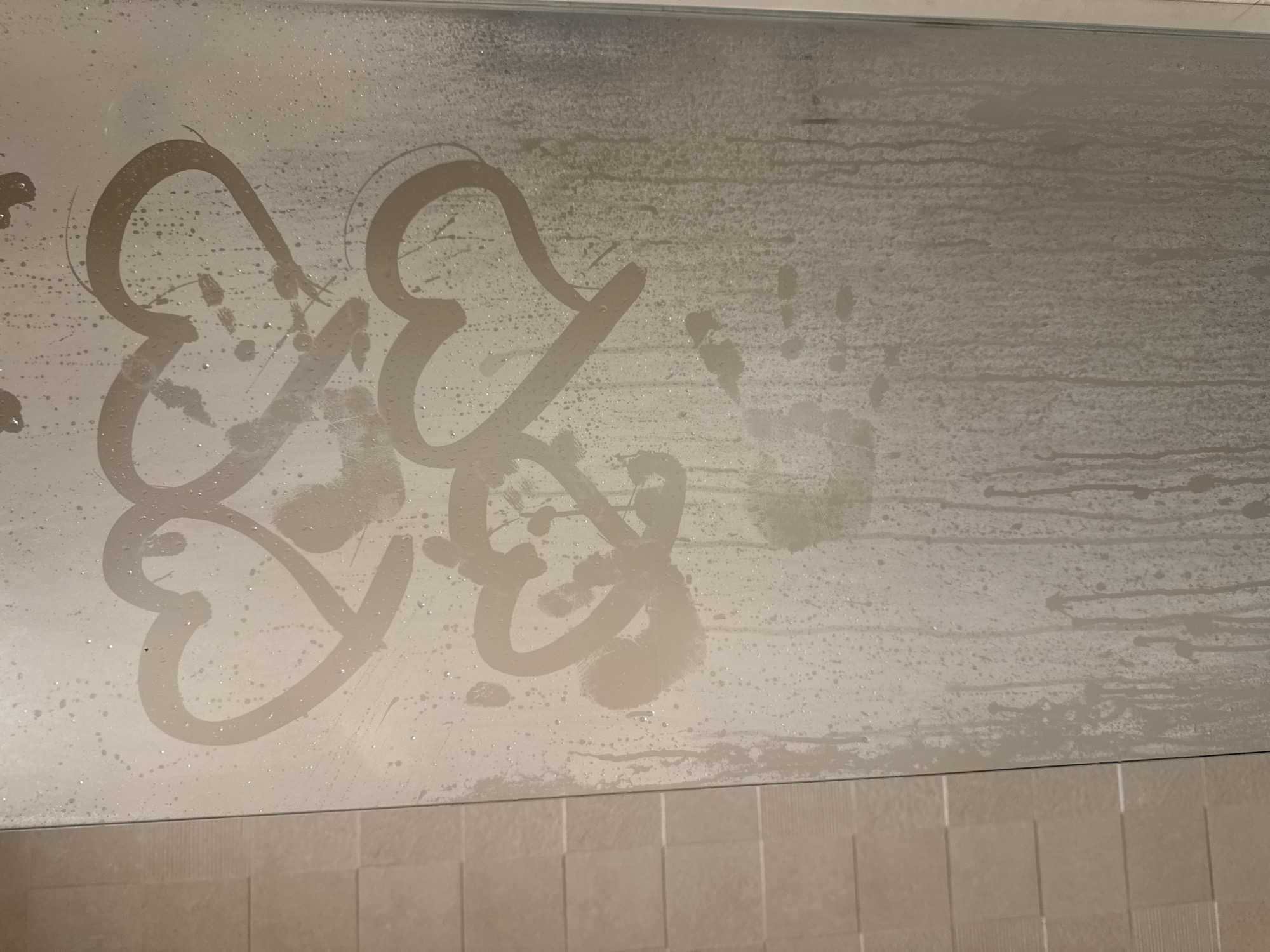The image captures a close-up view of a steamy glass surface, most likely a shower door or bathroom mirror, heavily fogged with condensation. Numerous small water droplets and larger streaks where moisture has run down are visible across the entire surface.
On the left side, three distinct heart shapes have been drawn into the condensation, clearing the fog to reveal the glass beneath. Two larger hearts appear intertwined or closely placed at the upper left, while a smaller, single heart is positioned below and slightly to their right. The outlines of these hearts are clear and stand out against the misty background.
To the right of the drawn hearts, a faint, smudged impression of what appears to be a handprint is visible in the condensation.
Along the very bottom edge of the frame, a segment of light-colored, possibly beige or cream, square tiles can be seen, providing a contextual cue that the setting is a bathroom. The tiles are uniform in size and arranged in a grid pattern.
The overall scene suggests recent use of a shower or bath, creating the warm, humid conditions that fogged the glass. The drawn hearts convey a sense of affection, playfulness, or personal touch. No visible text is present in the image.