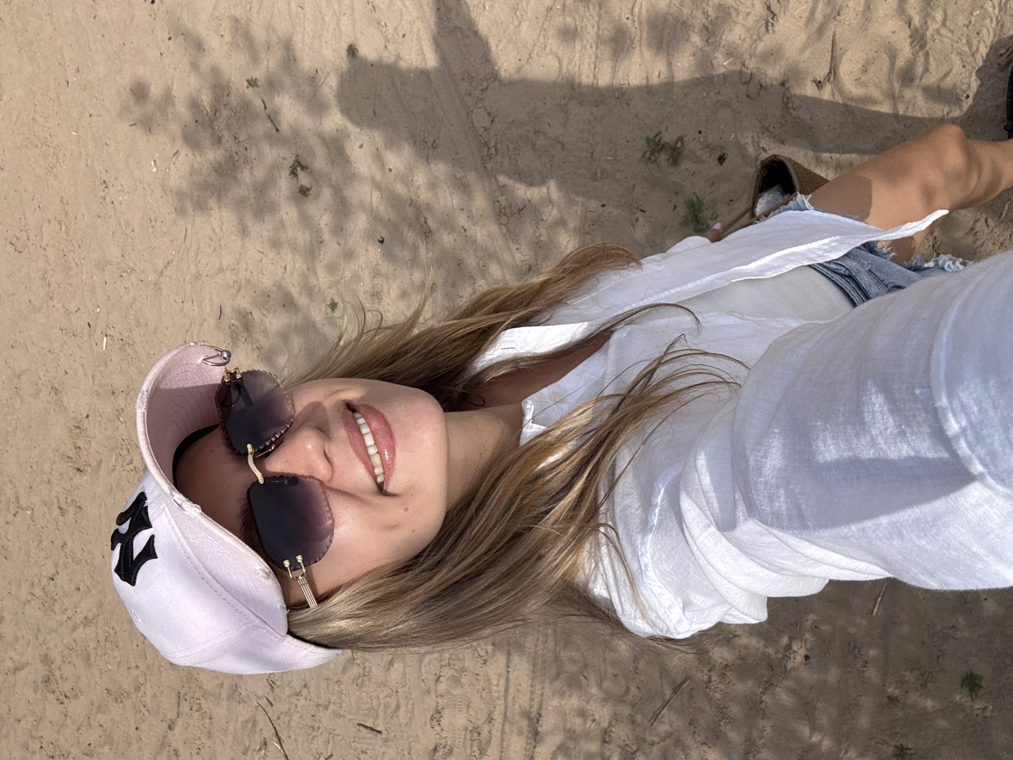 A young woman with long blonde hair is lying down on sandy ground, possibly a beach or park. She is wearing a white button-down shirt, denim shorts, and a light pink baseball cap with a black logo that appears to be a stylized letter 'N' or 'Y'. She is also wearing oversized sunglasses with dark lenses and gold accents. Her head is tilted back, and she is smiling broadly, showing her teeth. Her hair is spread out around her face and shoulders. The ground is a light brown sand with scattered dry leaves and twigs. Sunlight casts shadows from unseen objects onto the sand, suggesting it is daytime. The overall mood appears relaxed and happy.