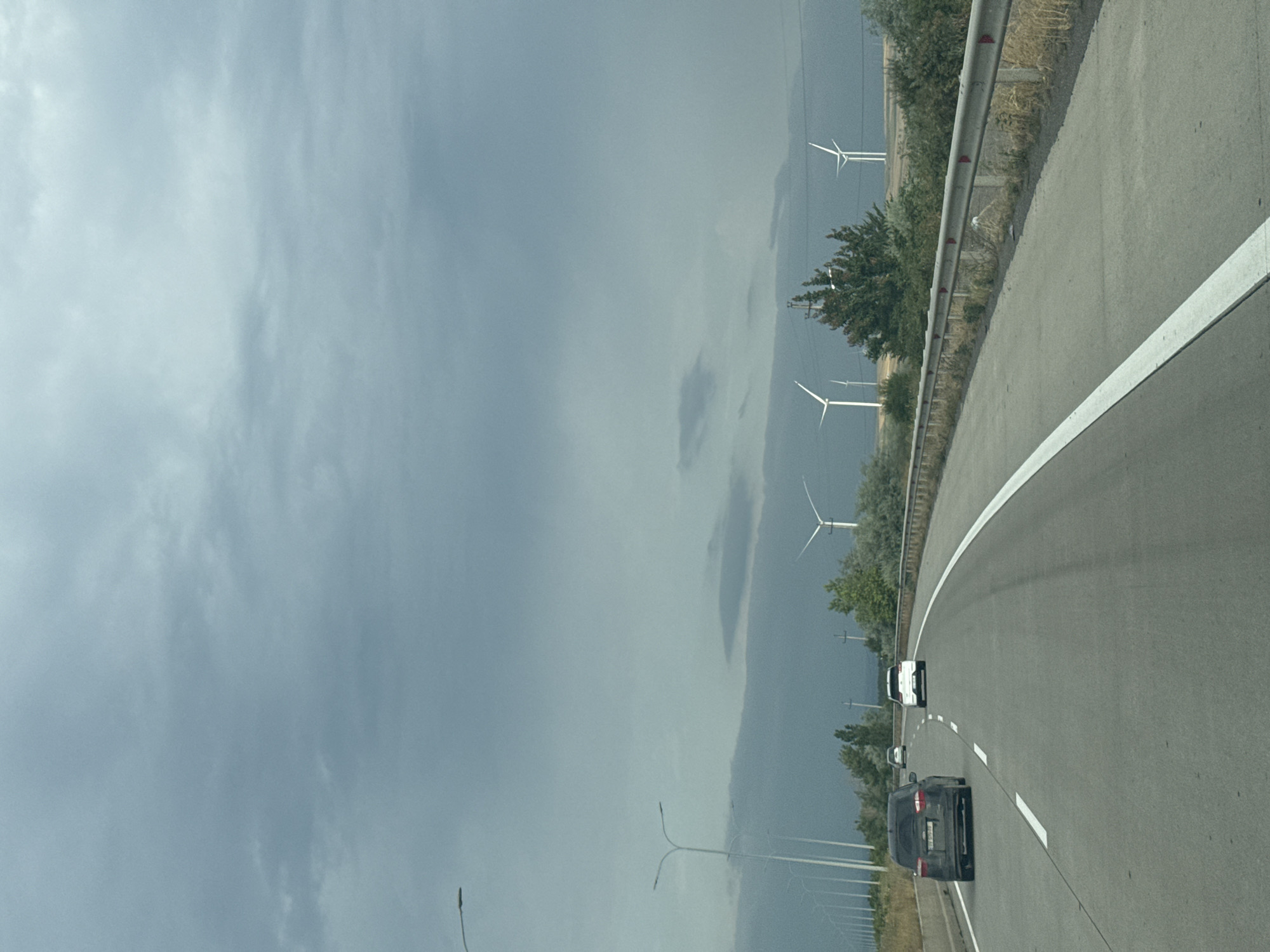 The image captures a highway scene with a cloudy, overcast sky. In the distance, several wind turbines are visible, suggesting an open, possibly rural area. The road is paved, with clear lane markings. On the road, there are several vehicles: a dark-colored car is in the left lane, and a white car is in the right lane ahead of it. Further up the road, another white car is visible. The roadside features vegetation, a guardrail, and streetlights along the left side. The overall atmosphere is muted due to the cloudy weather. There is no visual information to confirm the location is Skra, Georgia.