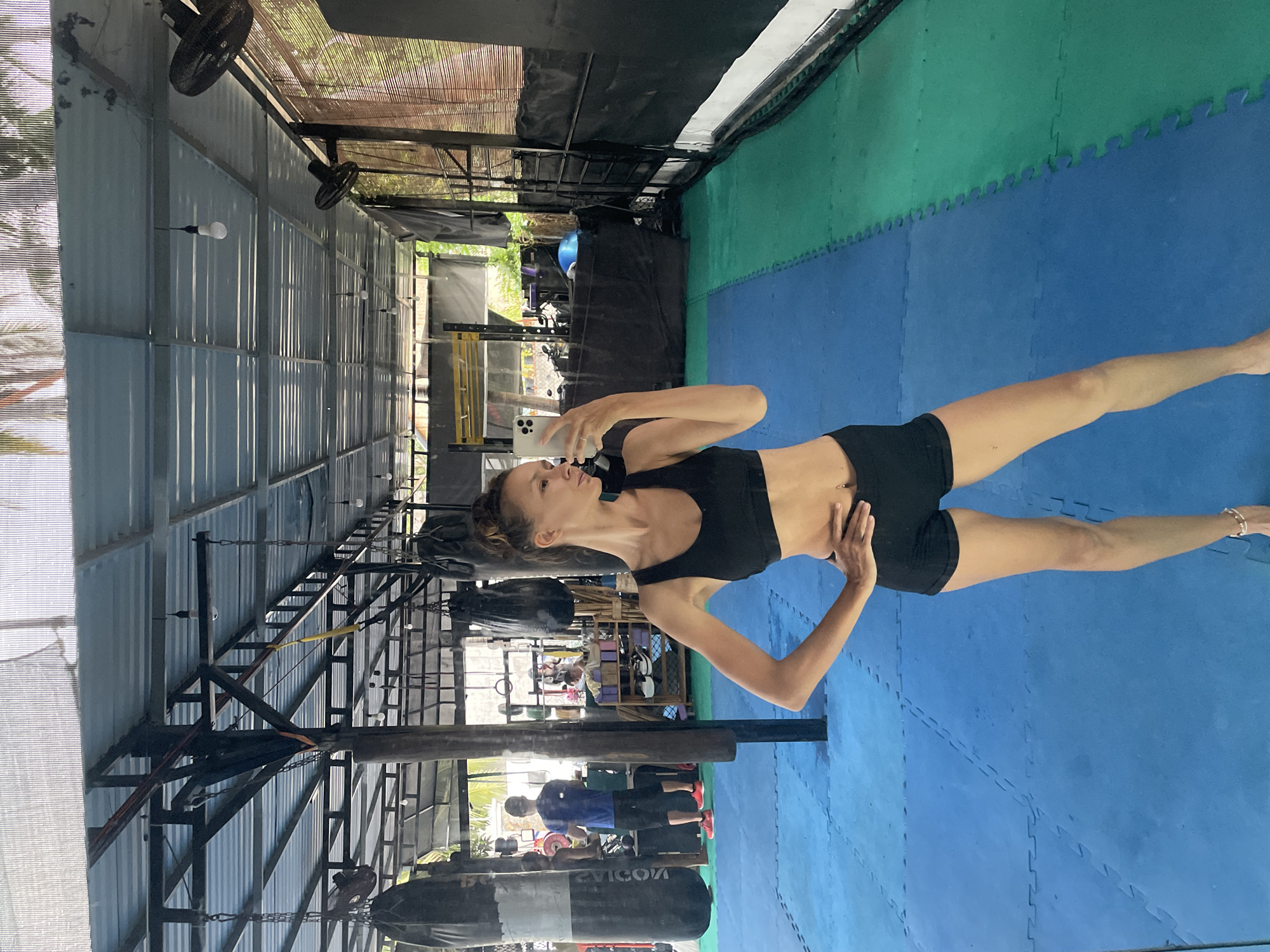 An image captures a woman posing in what appears to be an open-air gym or martial arts training facility during the daytime. She stands in the foreground, facing left, holding a white smartphone, likely taking a selfie or recording herself. She is dressed in a black sports bra and black shorts, with her hair pulled back, and has a navel piercing.
The floor of the facility is covered with interlocking blue and green exercise mats. To the woman's right, a large black punching bag hangs from the ceiling structure. In the background, two men are visible; one in a blue shirt stands near another punching bag, and another person is partially visible further back among various gym equipment such as weights and racks. The facility features a utilitarian construction with metal beams, corrugated metal walls, and a roof partially covered by bamboo-like screening and black fabric, providing shade. Multiple black ceiling fans and hanging light bulbs are also visible. Bright natural light illuminates the scene, suggesting a clear day. The word "EVVICOM" is partially visible in reverse on one of the punching bags. The setting reflects an active training environment in a warm climate.