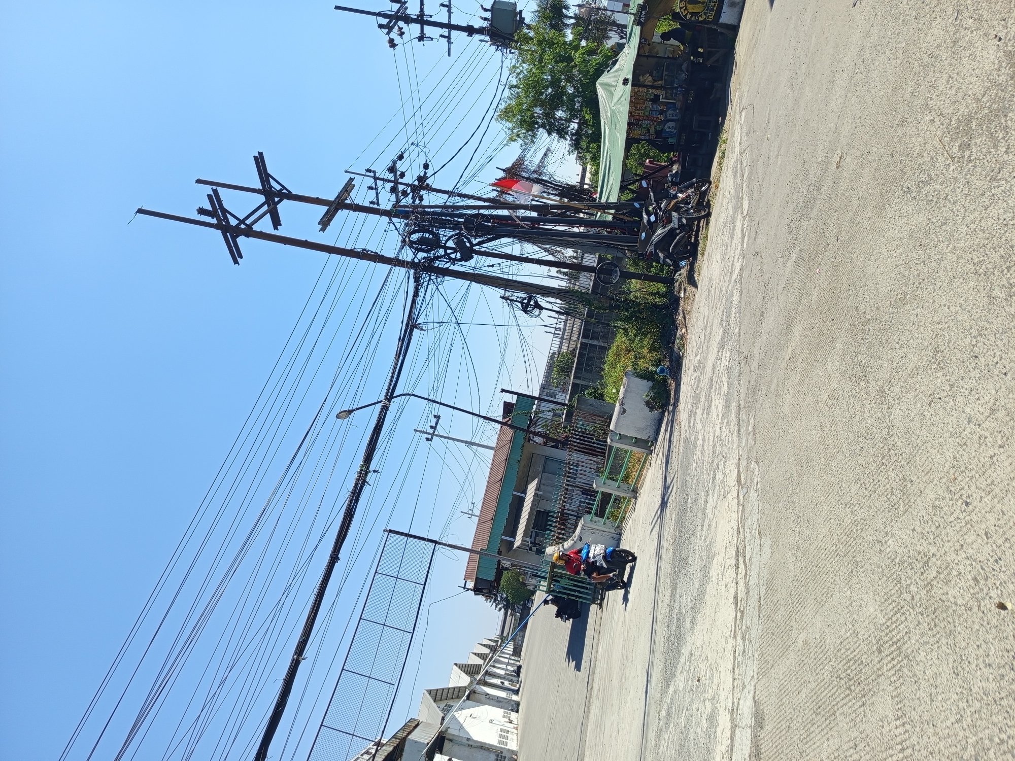 The image captures a scene in Sawotratap, Indonesia, on a bright, sunny day. The foreground shows a paved road with a textured surface. To the left, a white building with a long, sloped roofline and multiple window openings suggests an industrial or commercial facility. A person wearing a red shirt and yellow helmet rides a blue and white scooter towards the viewer, on the left side of the road. A second motorcycle is parked behind the scooter.
The middle ground features a row of poles supporting a complex network of overhead electrical wires. One of the poles displays a small Indonesian flag. Adjacent to the poles is a green awning stretched over a stall or small shop, where several people are gathered. Two motorcycles are parked near the stall.
The background is dominated by a clear blue sky. The overall environment appears to be an urban or semi-urban area with industrial structures, utility infrastructure, and a local market or roadside stall.