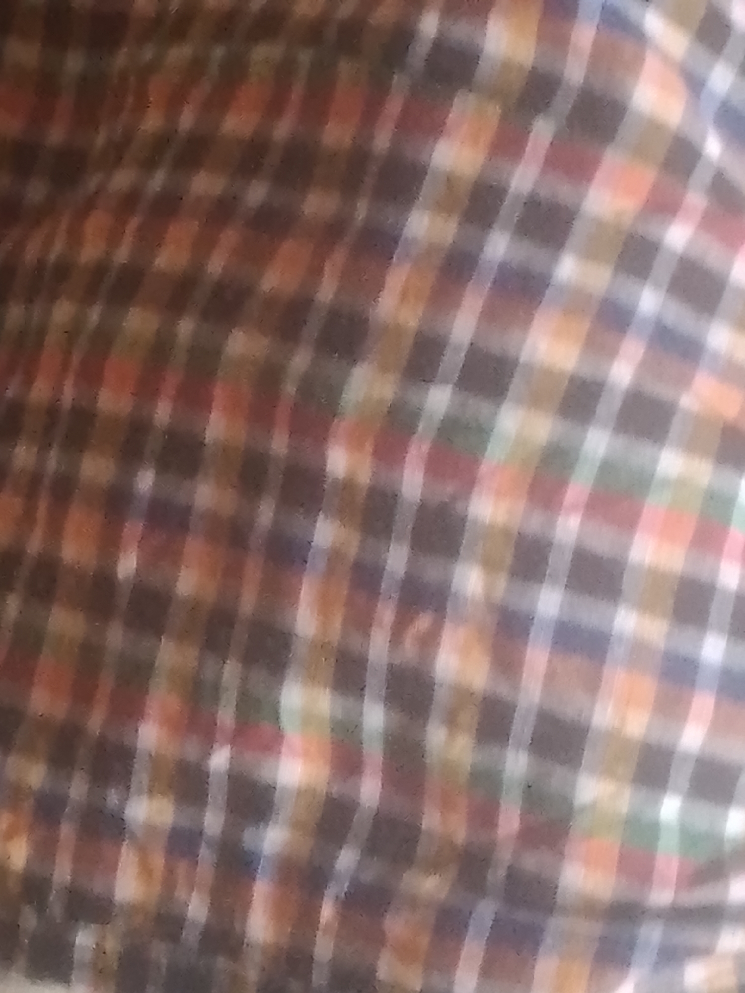 The image is a close-up, blurry shot of a plaid fabric. The pattern consists of intersecting lines of various colors, including brown, red, orange, white, green, and blue, forming a checkered design. The fabric appears to be slightly wrinkled and the lighting is dim, making it difficult to discern any specific details or textures. There is no discernible context for its location in Jauro Yinu, Nigeria, as the image only shows the fabric itself.