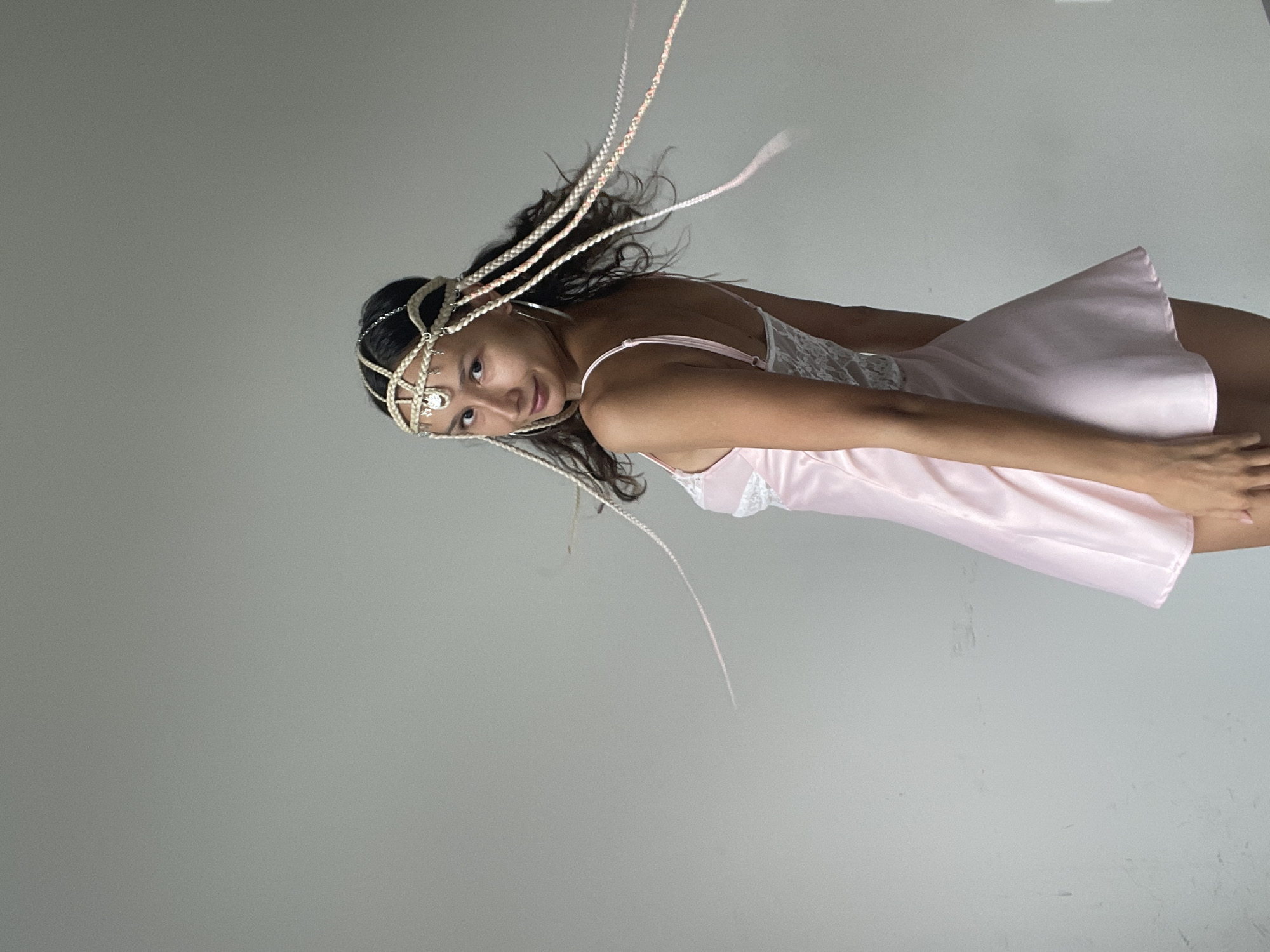 A young woman with long, dark hair braided with light-colored ribbons and adorned with a pearl and jewel headpiece stands against a plain, light gray wall. She is wearing a short, light pink slip dress with lace detailing on the bodice and sides. The dress is made of a satin-like material. Her hair is styled in braids that trail down her back and to the side, some appearing to be in motion, suggesting she may have turned. She is looking over her left shoulder at the camera with a slight smile. The image was taken indoors, likely in a studio or room with a seamless background, with no visible indication of it being in Mũi Né, Vietnam.