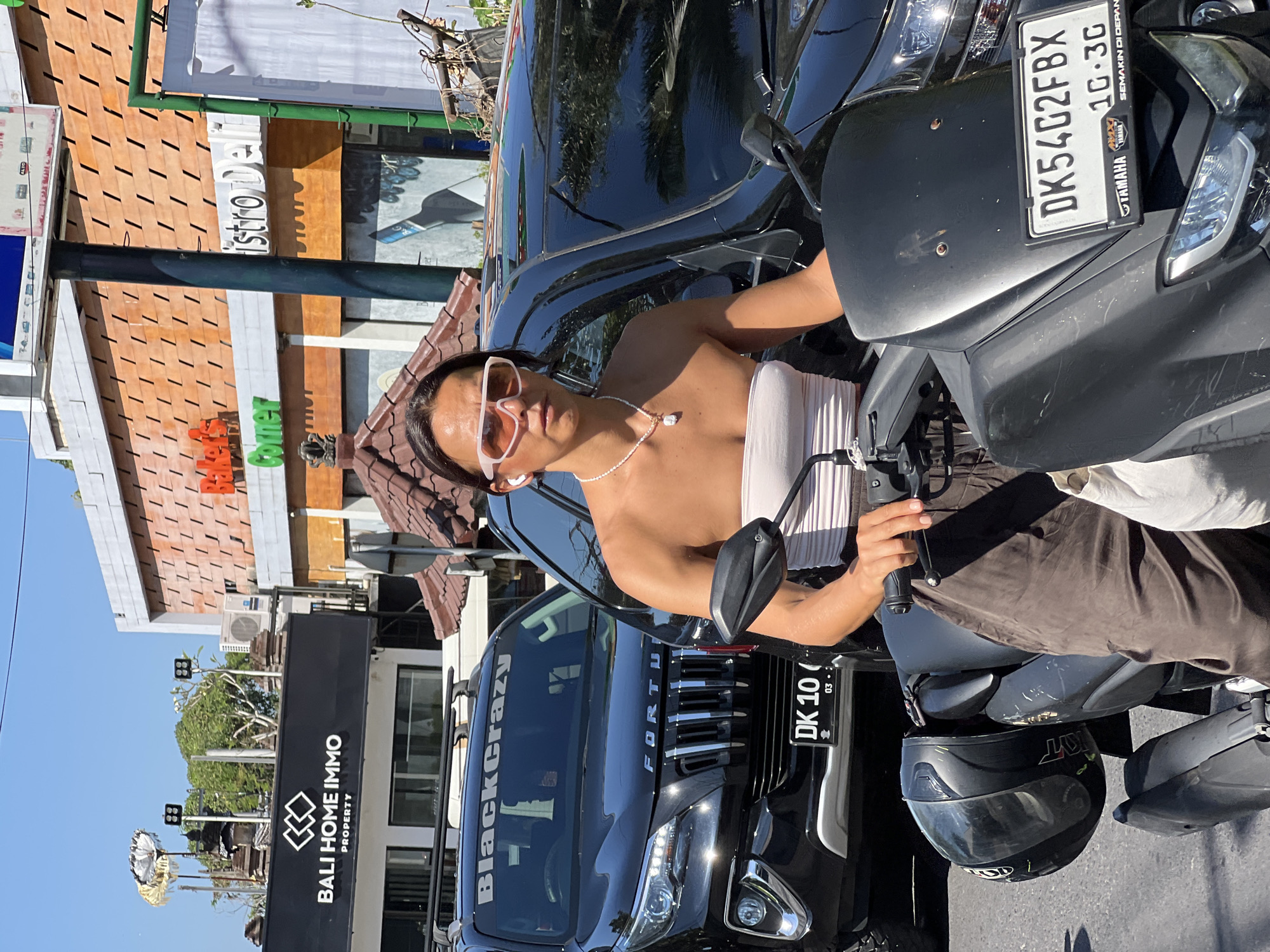 A person with dark hair and wearing sunglasses and a strapless white top is seated on a dark-colored scooter. The scooter has a black windshield and a license plate that reads "DK54G2FBX 10 3G". The person is wearing a pearl necklace with a pendant. To the left of the scooter, a black SUV is parked. The SUV has a license plate that reads "DK 10 03-". The windshield of the SUV has a sticker that says "BlackCrazy". In the background, there are buildings with signs that read "BALI HOME IMMO PROPERTY", "Baker's Corner", and "istro Deli". The scene appears to be in a city, possibly Ubud District, Indonesia, given the context provided. The weather is sunny.