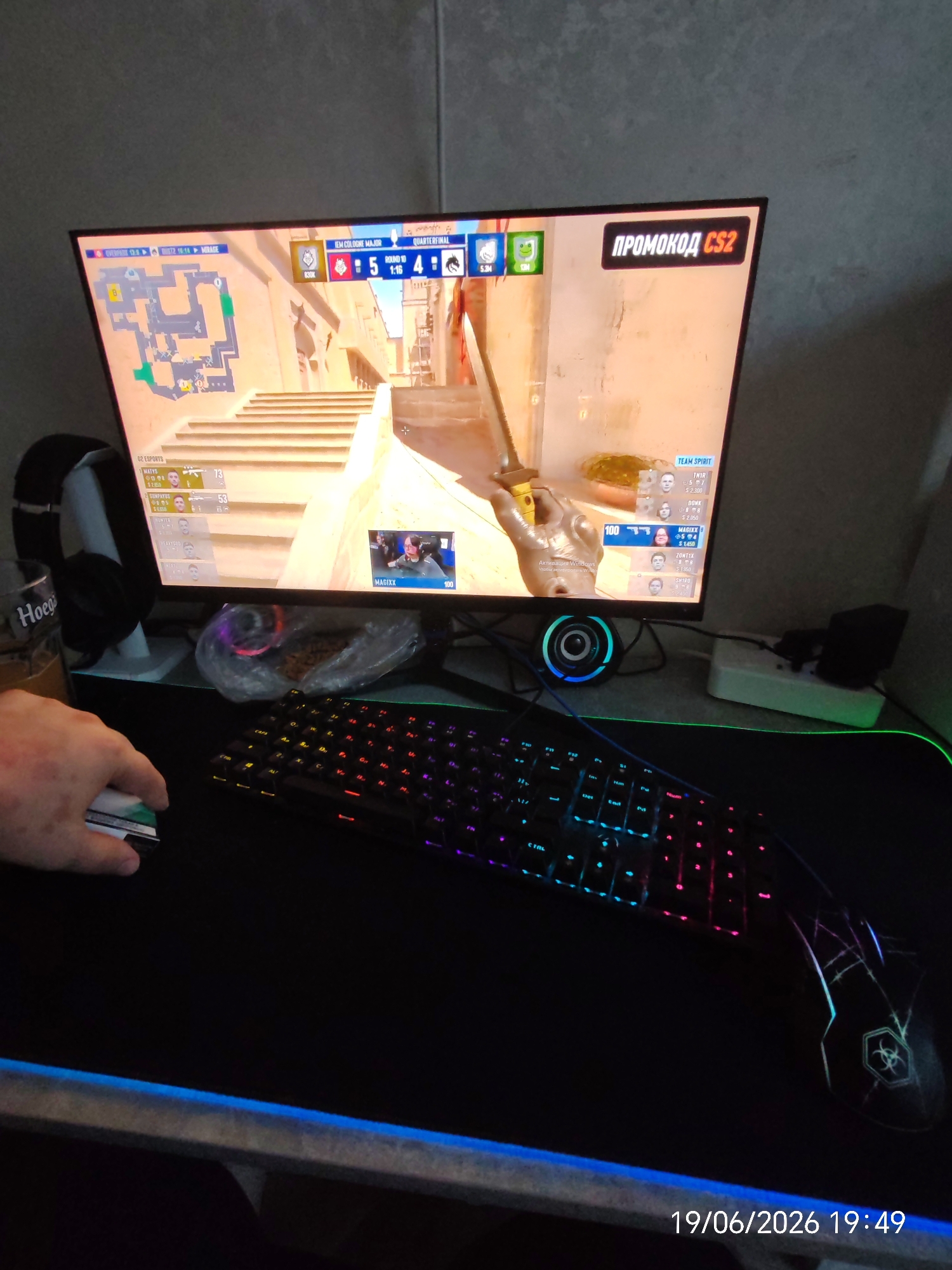 In an indoor setting, likely a room in Yablonovsky, Russia, a computer monitor displays a scene from a video game, possibly Counter-Strike 2, given the "PROMOCODE CS2" text. The game interface shows two teams, "TEAM SPIRIT" and "OG ESPORTS," with player names and scores. On the desk, a person's hand is visible holding a computer mouse. A colorful, illuminated keyboard is positioned in front of the monitor. Headphones rest on a stand to the left of the monitor, and a glass with branding partially visible as "Hoega" is also present. The desk is covered by a large mousepad with glowing blue trim. A power strip with plugs is visible on the desk to the right. The overall environment appears to be a gaming setup. The timestamp "19/06/2026 19:49" is overlaid on the bottom right of the image.