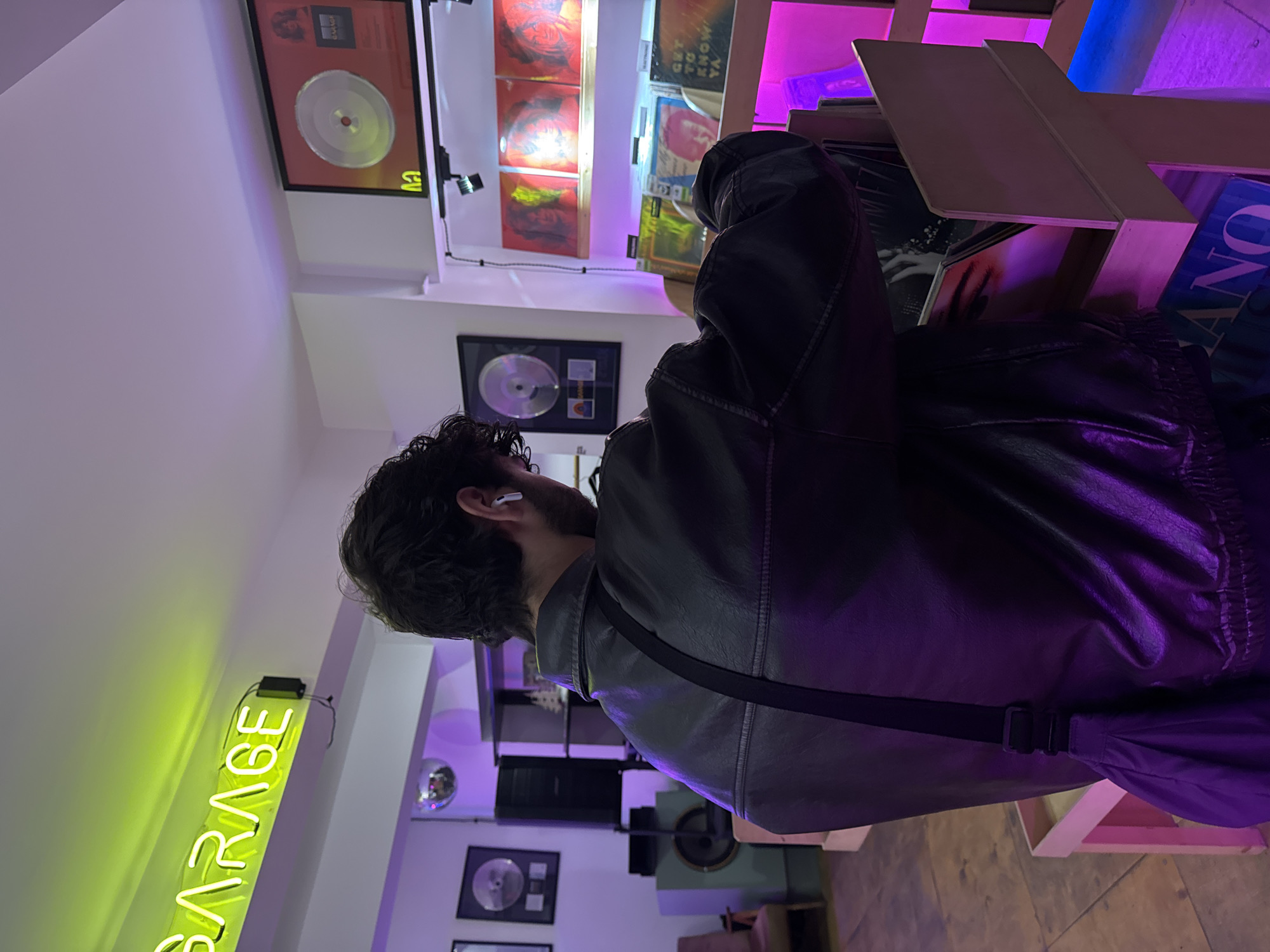 A vertically oriented photograph shows a man wearing a black leather jacket and an earpiece, browsing records at a vinyl record store. The store appears to be in Yerevan, Armenia, based on the "GARAGE" sign, a popular Yerevan music store. The man is standing in front of a shelf of records, with several vinyl albums visible. Behind him are framed gold records mounted on the wall. The store is illuminated with neon lighting, with the "GARAGE" sign in green and accent lighting in pink and purple. The man is wearing a black strap across his chest, possibly a shoulder bag.