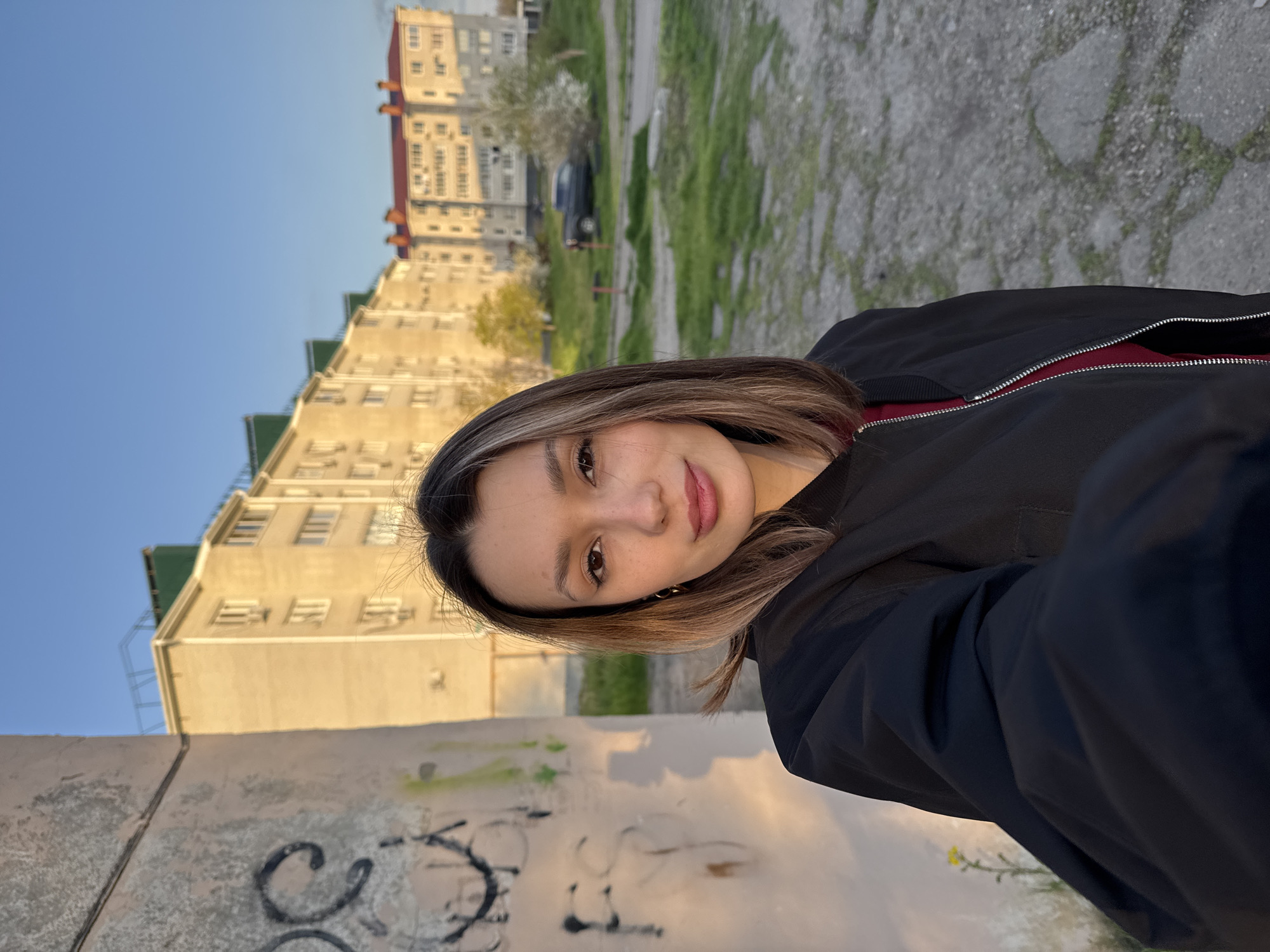 A young woman is featured in a close-up selfie taken outdoors, positioned on the right side of the frame and looking directly at the camera with a neutral, slightly smiling expression. She has medium-length brown hair with lighter highlights. She is wearing a black bomber-style jacket with a visible red lining or undergarment beneath. Small gold hoop earrings are visible.
The setting appears to be an urban or suburban residential area during the golden hour, likely late afternoon or early evening, indicated by the warm, directional sunlight that illuminates parts of the buildings and the woman. The sky is clear and blue.
In the background, on the left side of the image, multiple multi-story residential buildings with numerous windows and some green/red roof sections are visible. A dark-colored car is parked on a patch of grassy and unpaved ground between the buildings and the foreground. The ground further back consists of a mix of grass, dirt, and potentially broken pavement.
In the immediate foreground, a textured concrete or stone wall is prominent, covered with light-colored graffiti that includes abstract shapes and some partially discernible letters, such as "SC" and other squiggles. The ground in the foreground beneath the woman and the wall is paved with large, irregular grey stones or concrete slabs. A single small yellow flower is visible near the bottom left corner of the frame.
