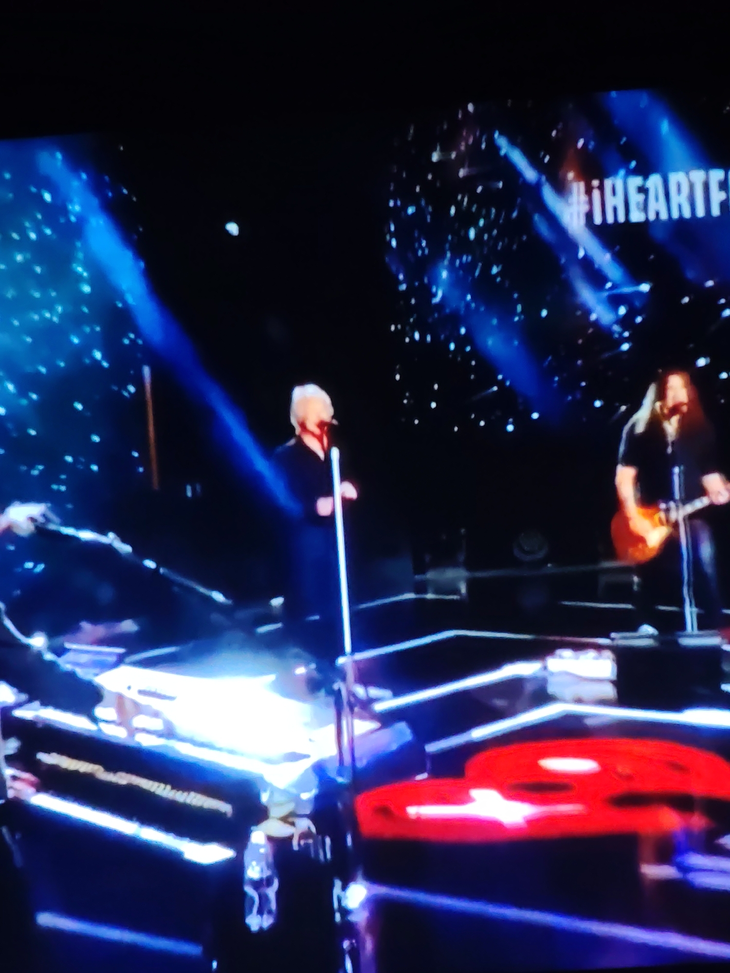The image shows a dimly lit stage with a blue and white light show. On the stage, there are three people. On the left, a person is partially visible, seated at a keyboard. In the center, a person with short, light-colored hair stands behind a microphone on a stand, wearing a dark outfit. To the right, a person with long, dark hair plays an electric guitar, also wearing a dark outfit. A hashtag "#iHEARTF" is visible in white lettering against the dark background on the right side of the image. The overall scene appears to be a live performance or broadcast. There is no information within the image to indicate a location in Vladimir, Russia.