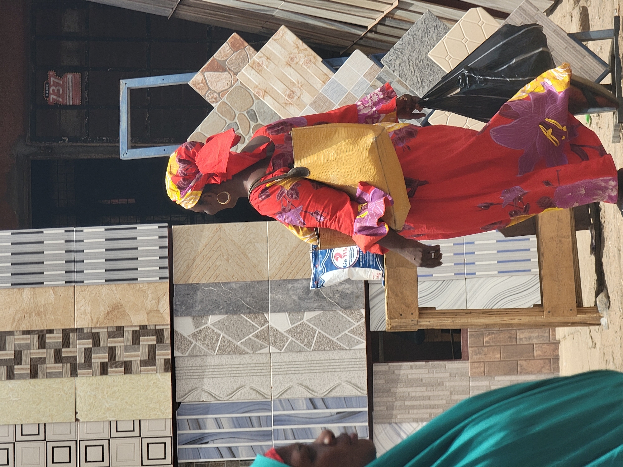 In Maiburiji, Nigeria, an African woman wearing a red and yellow head covering and a vibrant red dress with purple floral patterns stands amidst displays of ceramic tiles. She carries a yellow purse. Adjacent to her, different patterns and styles of tiles are arranged, presenting options for flooring or wall decor. The setting suggests a commercial area, specifically a shop or market specializing in building materials. Another person, partially visible in the foreground, is wearing a green garment.