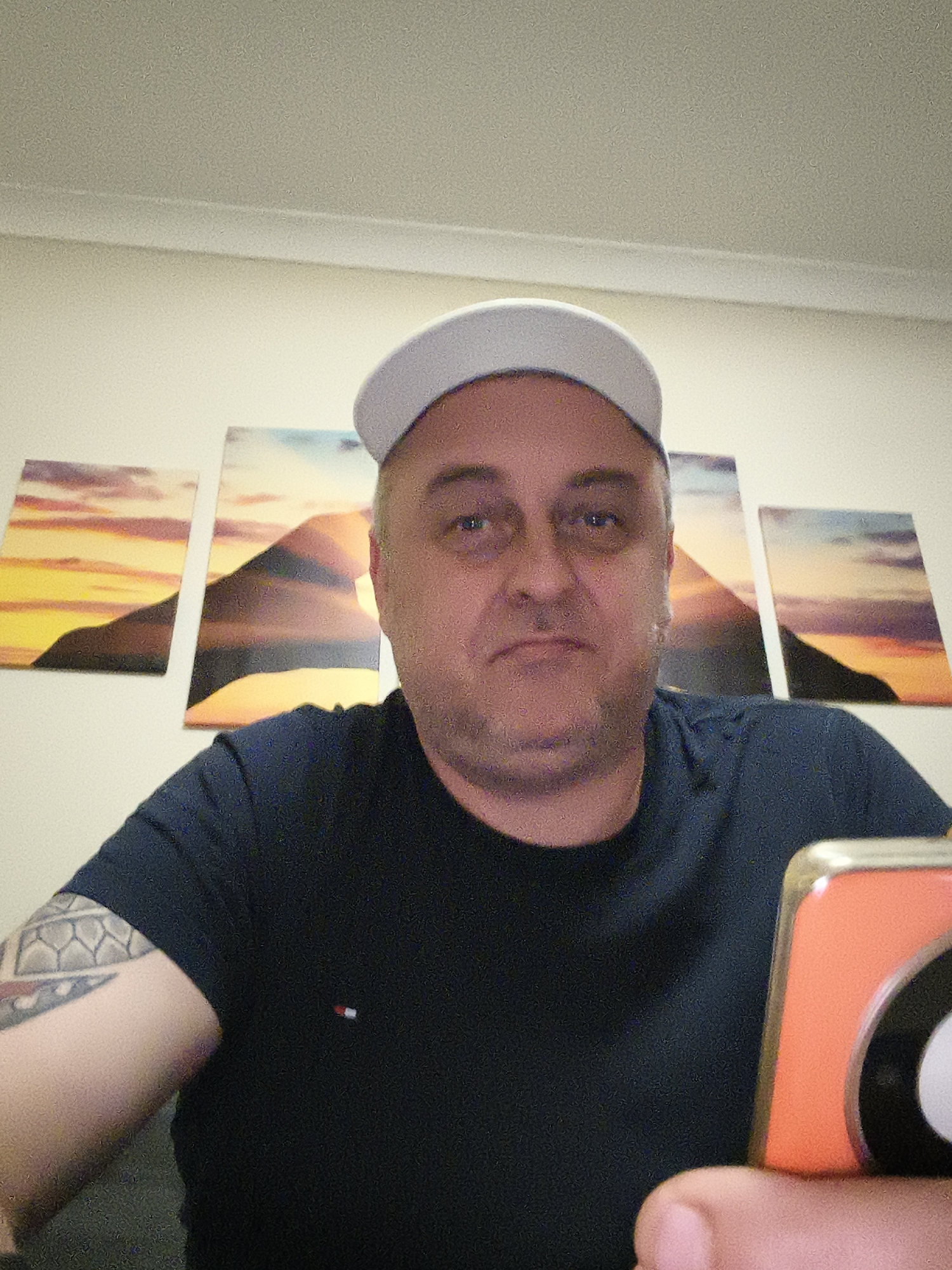 A man, likely middle-aged, is pictured indoors in a medium close-up, looking directly at the camera with a neutral expression. He wears a light-colored baseball cap and a dark t-shirt with a small red, white, and blue embroidered logo on the left chest, resembling the Tommy Hilfiger brand. He has stubble on his face and a visible tattoo with a geometric or tribal pattern on his left bicep. In his right hand, partially visible in the bottom right corner, he holds a bright orange smartphone with a distinctive black and white camera module.
Behind him, on a light-colored wall, are four horizontally arranged rectangular canvas prints. These prints depict a continuous panoramic landscape scene of mountains or hills silhouetted against a vibrant sunset or sunrise, rendered in warm yellows, oranges, and purples. A white ceiling and molding are visible at the top of the frame. The setting suggests a domestic interior, consistent with a residence in Huntingdonshire, United Kingdom. The man appears to be posing for a photograph or taking a selfie.