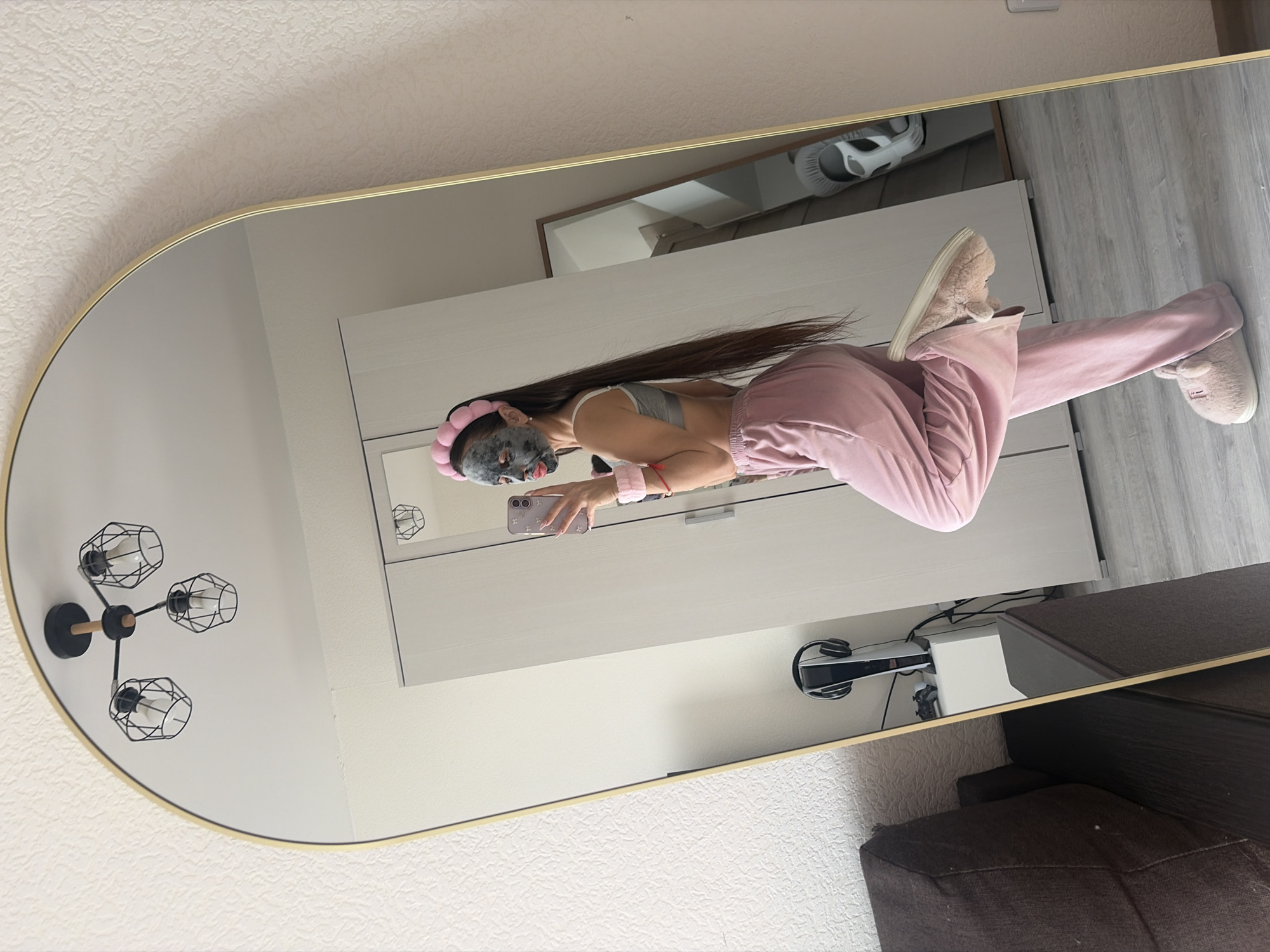 This media file is a mirror selfie taken indoors, rotated 90 degrees counter-clockwise. The main subject is a person, likely female given the long hair, standing in front of a large, gold-framed, oval-shaped mirror. The person is wearing a grey sports bra, pink sweatpants, and fluffy light pink slippers. A dark-colored face mask covers their face, and a pink, plush headband holds their long, dark hair back. They hold a light-colored smartphone with their left hand, capturing their reflection. A red bracelet is visible on their left wrist.
The setting appears to be a modern residential room in Saint Petersburg, Russia, likely a bedroom or living area. Reflected in the mirror behind the person is a light-colored wardrobe or cabinet with a silver handle. On a surface beside the wardrobe, a white PlayStation 5 console and a pair of white headphones or a PlayStation VR headset are visible, with wires tangled behind them. In the upper left corner of the mirror's reflection, a contemporary black ceiling light fixture with three exposed bulb cages is seen. The walls of the room have a light-colored, textured wallpaper, and the floor, visible in the reflection, is light-toned laminate or wood. A dark brown sofa or armchair is partially reflected in the lower right portion of the mirror.
The activity depicted is a self-care routine, with the person applying a face mask, and simultaneously taking a casual mirror selfie. The casual attire suggests it is either daytime or evening in a relaxed home environment. No discernible text is visible on any objects.