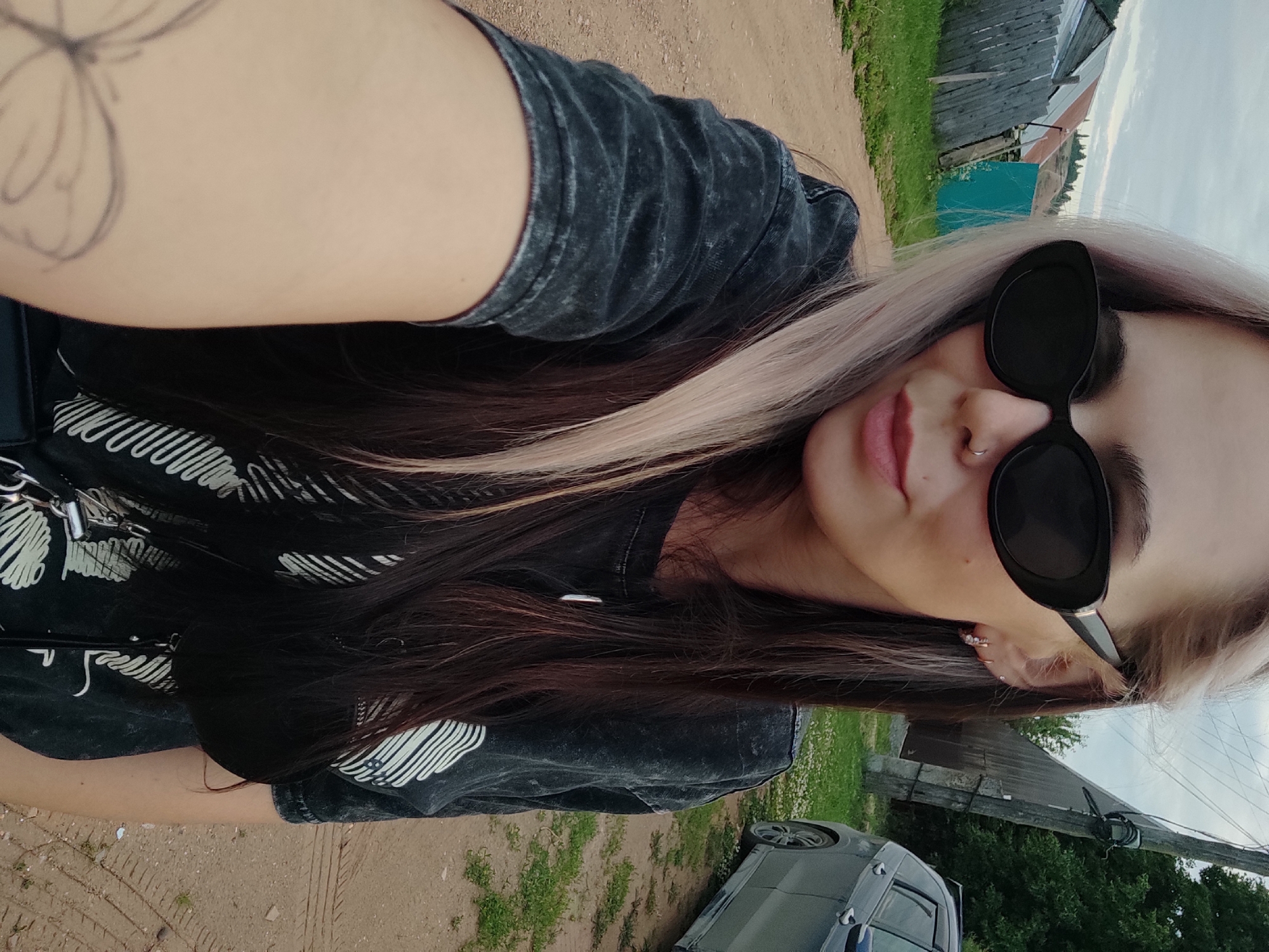 The media file is a vertical, close-up, selfie-style shot of a young woman with blonde and dark hair. She is wearing a dark t-shirt with a white graphic print, sunglasses, and has a nose ring and earrings. Her left arm is visible in the foreground, showing a butterfly tattoo. She is outdoors in what appears to be a rural setting, with a dirt road and some greenery visible. In the background, there are wooden fences, some buildings, and a parked car. The sky is cloudy, suggesting it might be overcast. The overall mood of the image is casual and relaxed. The location is identified as Olkhovka, Russia. There is no visible text in the image.