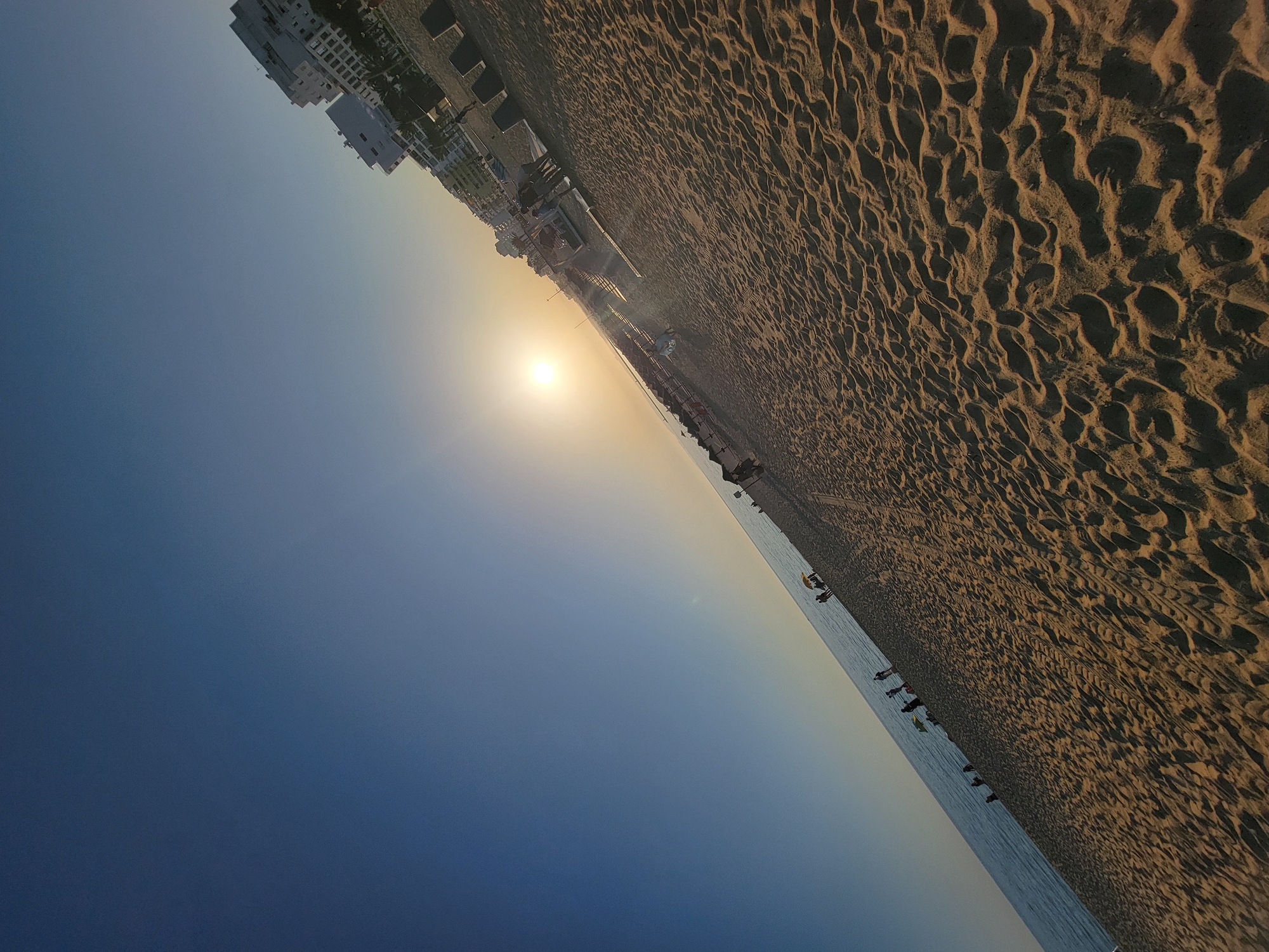 This is a wide-angle, tilted, eye-level shot of Quarteira beach in Portugal, during sunset. The scene is dominated by the sandy beach in the foreground, with the ocean to the left and buildings to the right. The sun is low on the horizon, casting a warm glow across the scene.
In the foreground, the sand is textured with ripples and shadows, indicating it is slightly damp. Towards the middle ground, there are numerous beach chairs and umbrellas, some of which are set up in rows. Small groups of people are scattered across the beach, some walking, some standing, and some sitting. A few individuals are visible near the water's edge, and one person is standing under a yellow umbrella.
Further back, along the coastline, a line of modern white buildings rises, interspersed with some palm trees and other vegetation. The buildings appear to be hotels or apartment complexes. Beyond the buildings, the sky transitions from a clear, deep blue at the top to a pale, almost white hue near the horizon, where the sun is visible as a bright disc. The ocean is a calm expanse of blue water, meeting the sand at a gentle angle. The overall impression is one of a peaceful, late afternoon on a popular European beach.
