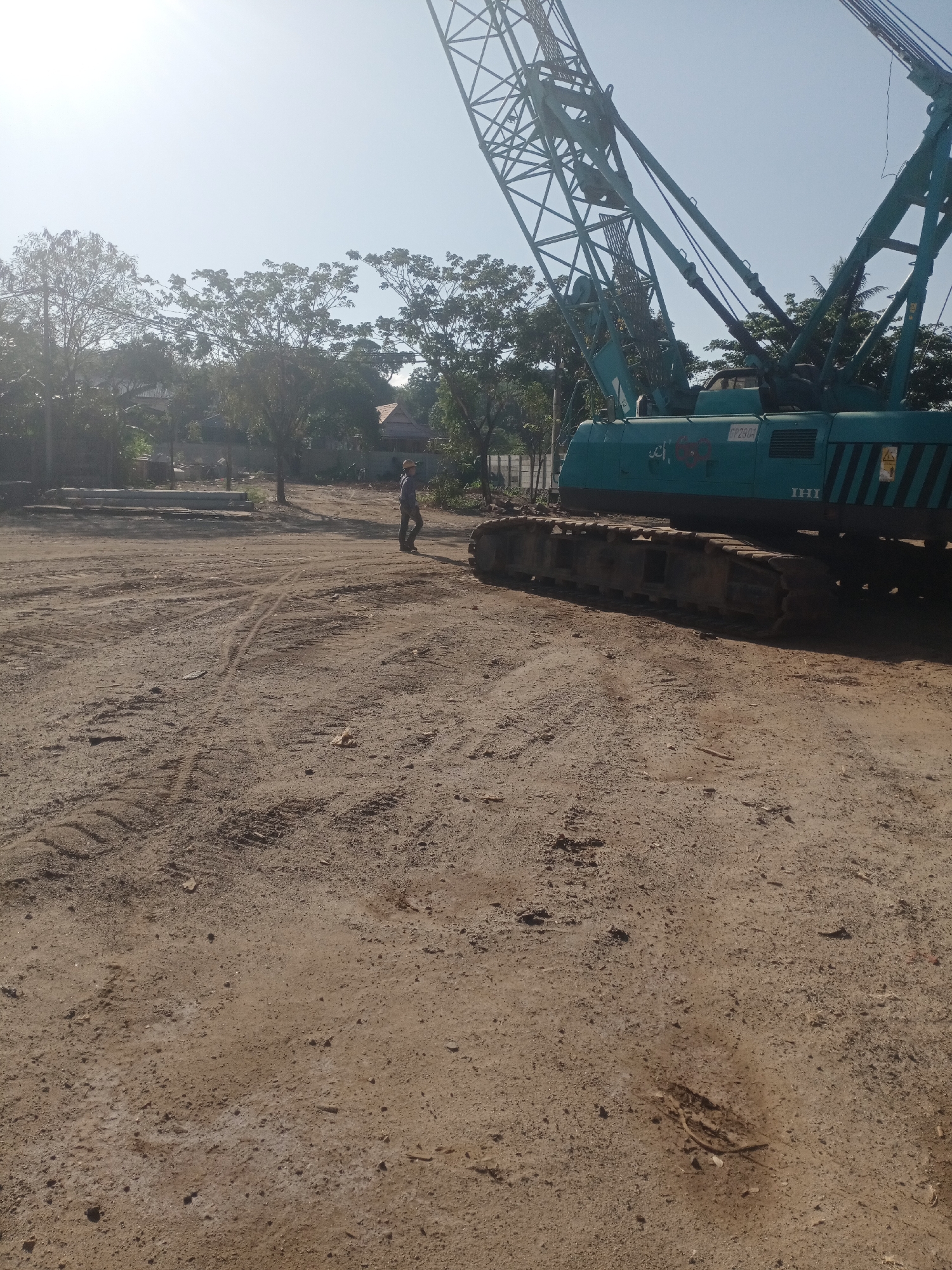 In the city of Siddo, Indonesia, a construction site is visible under a bright, hazy sky. The foreground is dominated by a dirt and gravel ground with tire tracks and scattered debris. To the right, a large, teal-colored crawler crane with a prominent lattice boom is parked. A person wearing a hat and dark clothing stands near the center of the frame, observing the scene. In the background, trees, some with sparse foliage, line the perimeter. A low concrete wall and what appears to be building materials are also present. The overall impression is of an active construction zone in a semi-arid environment.