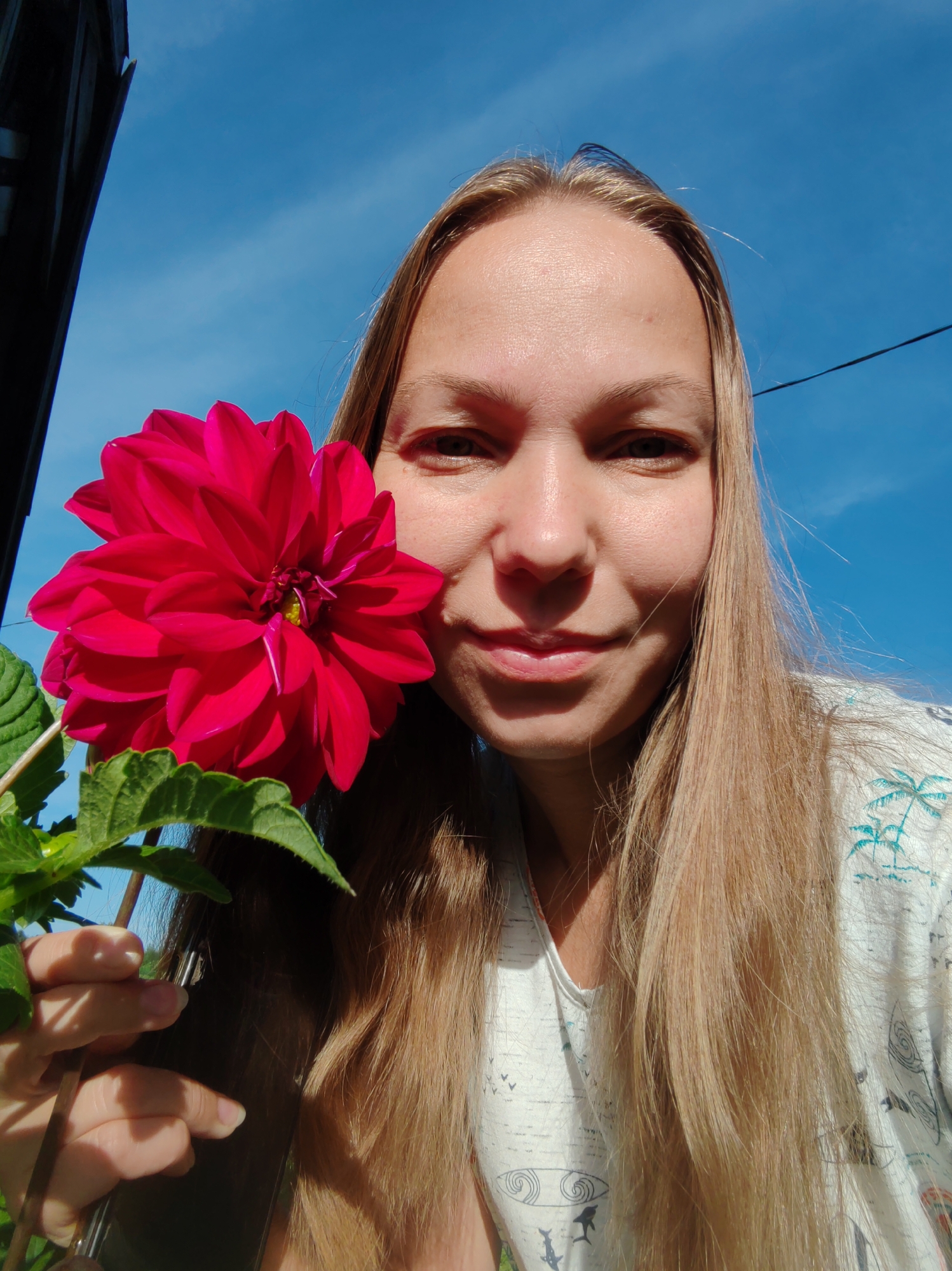 A blonde woman with long hair is taking a selfie outdoors, holding a large pink flower with green leaves. She is wearing a white t-shirt with a tropical print. The background is a bright blue sky with some faint clouds. The woman's face is partially obscured by the flower, but she is smiling. The image appears to have been taken during the daytime, with the sun casting shadows on her face. The location in the city of Чухлэм, Russia, is not discernible from the image.