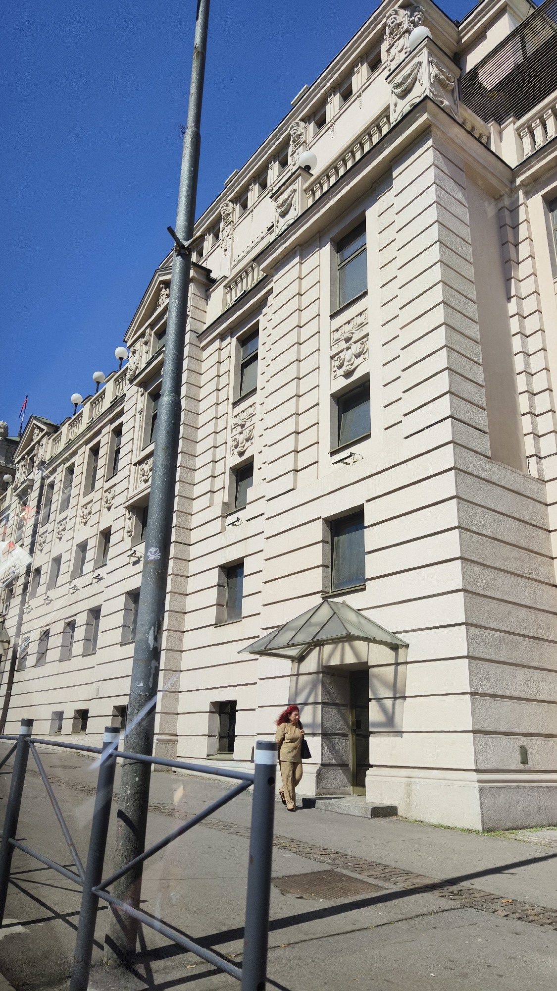 A medium shot shows a woman walking on a sidewalk in front of a light-colored building. The woman is wearing a tan-colored outfit and has red hair. She is carrying a black bag in her right hand. The building has a light-colored facade with many windows and decorative architectural details. A glass awning is visible over an entrance to the building. A metal pole and fence are in the foreground. The sky is clear and blue. The ground is paved with concrete and cobblestones. The scene appears to be a sunny day.