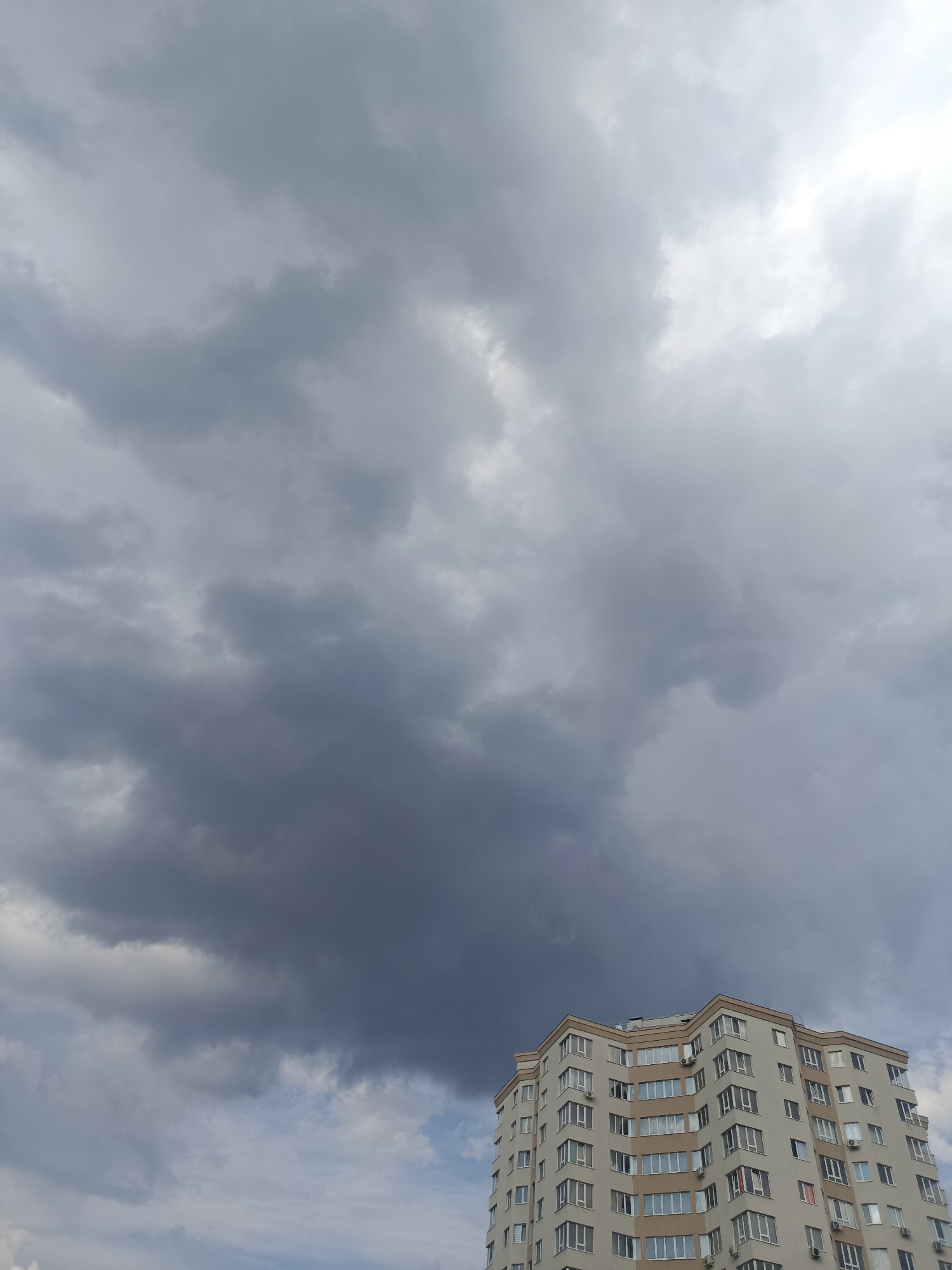 A multi-story apartment building with a beige and white facade stands against a dramatic sky filled with dark, heavy clouds. The building has numerous windows, some with red-tinted panes. The sky is a mix of dark gray storm clouds and patches of lighter sky, with a bright area suggesting the sun is either obscured or on the horizon. The image is taken from a low angle, looking up at the building and sky. This is a view of the residential building on Socoleni Street in Chișinău, Moldova.