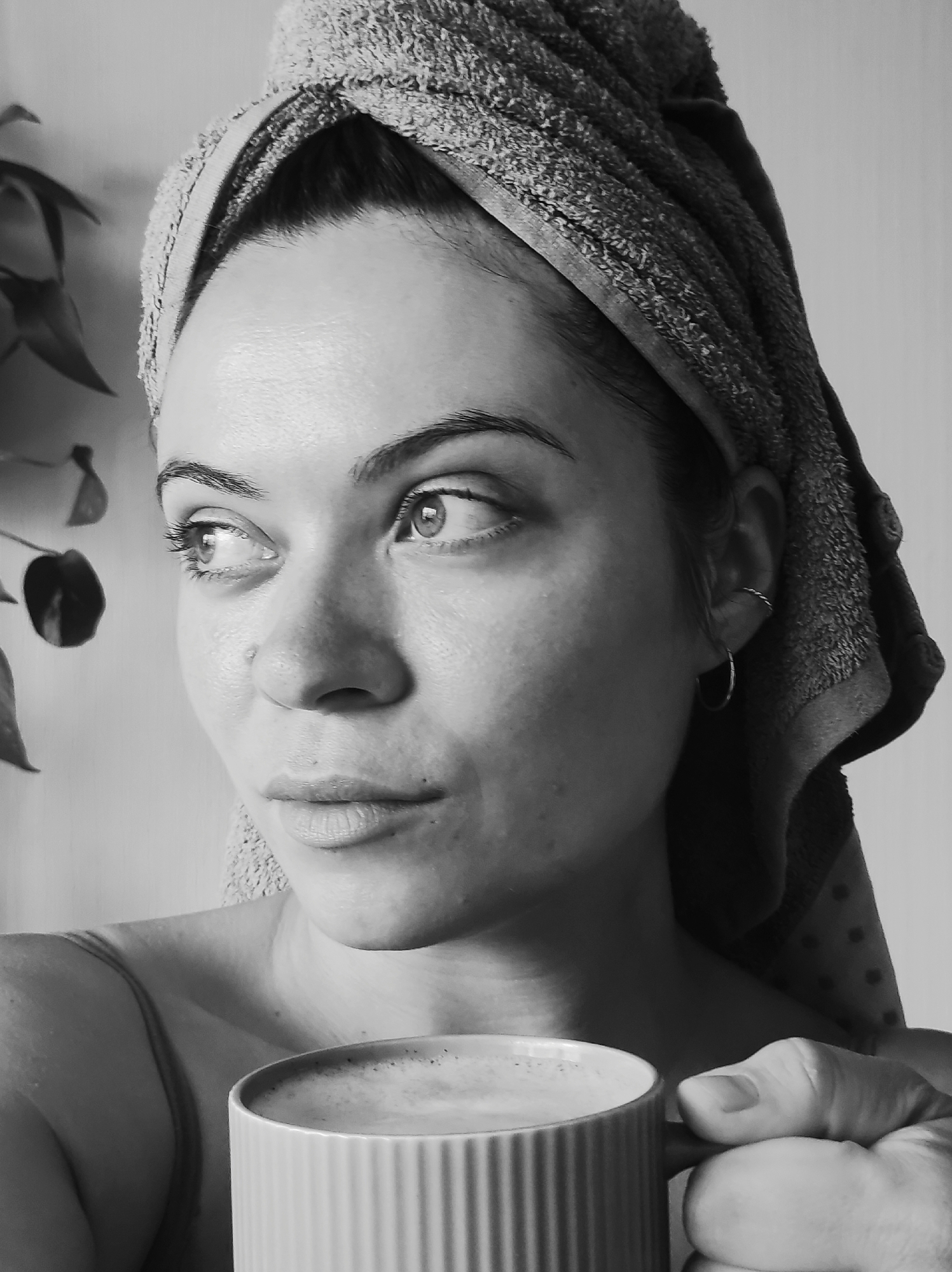 The black and white photograph shows a close-up, profile view of a woman with a towel wrapped around her head, holding a mug of coffee. She has a pensive expression and is looking off to her left. A plant is visible in the background on the left side of the frame. The woman is wearing a simple strap top. The lighting suggests an indoor setting, possibly a home, with natural light coming from the right. There is no indication of a specific location in Chaikovsky, Russia.