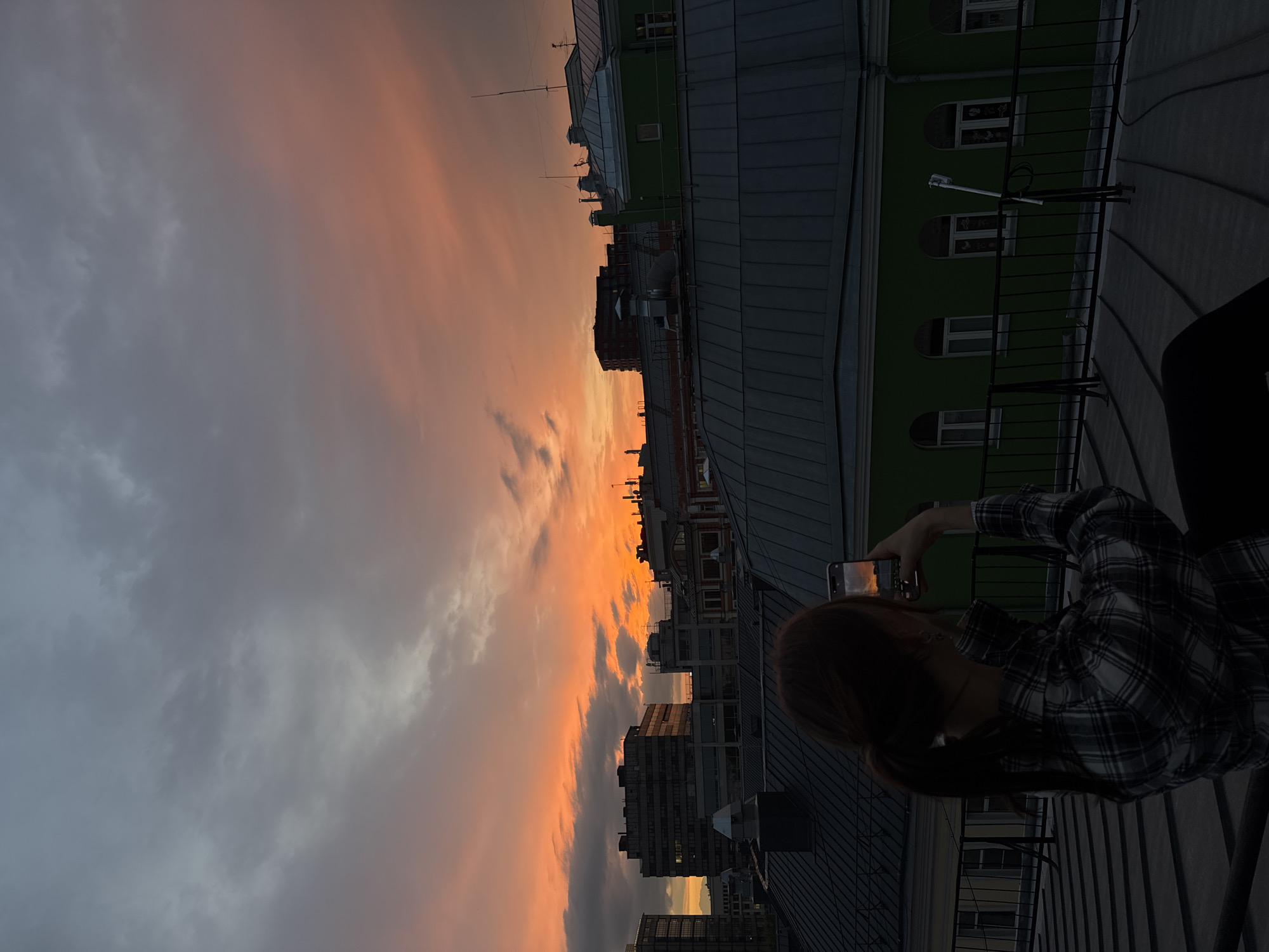 A young person is taking a photograph of a sunset in Moscow, Russia. The sunset colors the sky with shades of orange and gray. Tall buildings are visible in the distance, and the scene is set from a rooftop or elevated position. The person is wearing a plaid shirt. A phone is being held up in front of the person, capturing the sunset. The image is tilted, with the sky taking up the left portion of the frame and the buildings on the right.