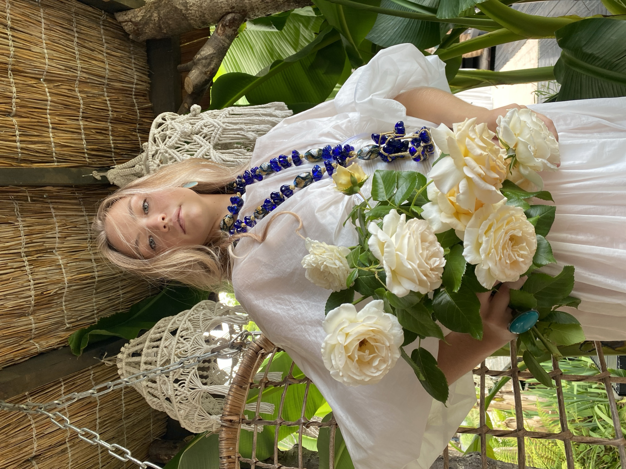 A young woman with blonde hair is posing outdoors, holding a bouquet of cream-colored roses. She is wearing a flowing white dress and a large, ornate blue necklace. The setting appears to be a tropical or subtropical environment, with lush green foliage and a thatched roof overhead, suggesting a location in Sochi, Russia, known for its subtropical climate. There are also macrame decorations hanging from the ceiling and a woven chair in the background. The woman is looking directly at the camera with a neutral expression.