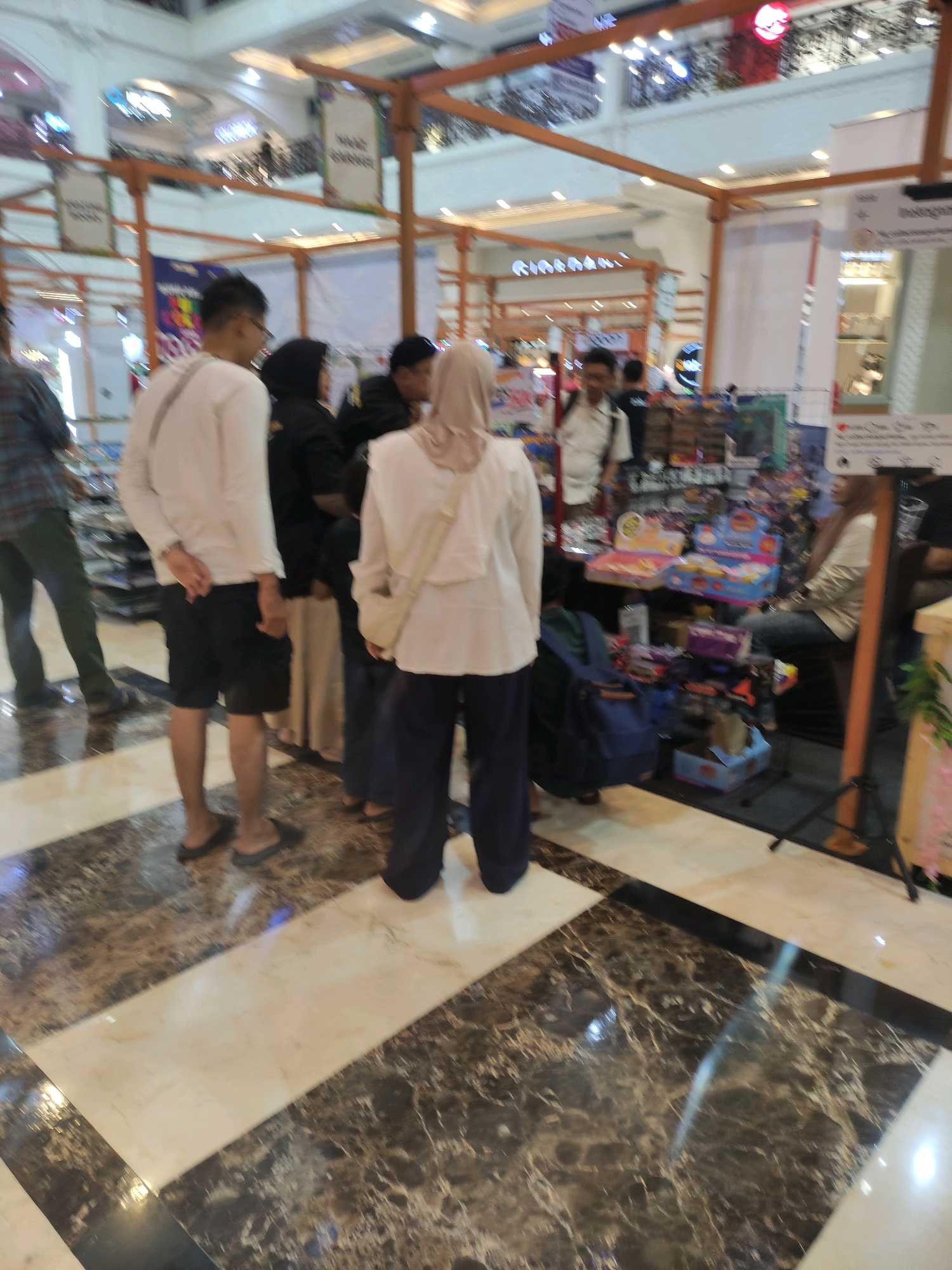 The image captures a busy scene inside a shopping mall, likely in Sinduadi, Indonesia, given the context. In the foreground, a man wearing a white long-sleeved shirt and black shorts stands with his hands behind his back, facing away from the camera. Next to him, a person in a black hijab and a woman in a light-colored hijab and a white top are also visible, with the woman in the white top facing away from the camera. In the background, more individuals are present, including a person in a plaid shirt and green pants walking left. Several stalls are set up, displaying various items, including what appear to be toys and packaged goods. The mall interior features large pillars and banners, with tiered levels visible in the distance. The floor is a polished marble with a distinctive pattern of dark brown and beige tiles. The lighting suggests an indoor environment with artificial illumination.