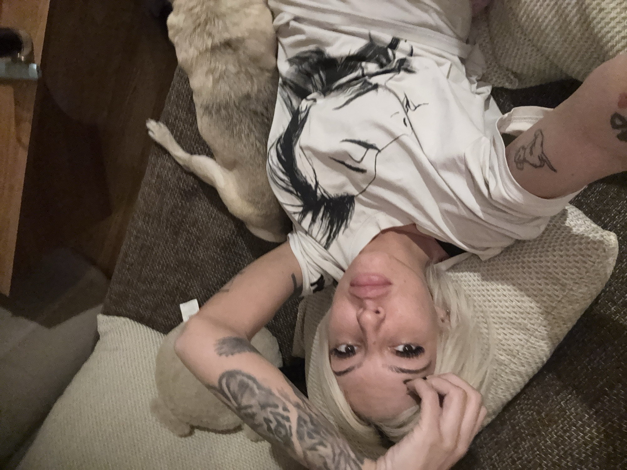 A woman is lying on a couch with her head on a pillow. A dog is lying on the couch next to her. The woman is wearing a white t-shirt with a black design on it. She has tattoos on her arms. The couch is brown. A wooden object is partially visible in the top left. Location is unknown.