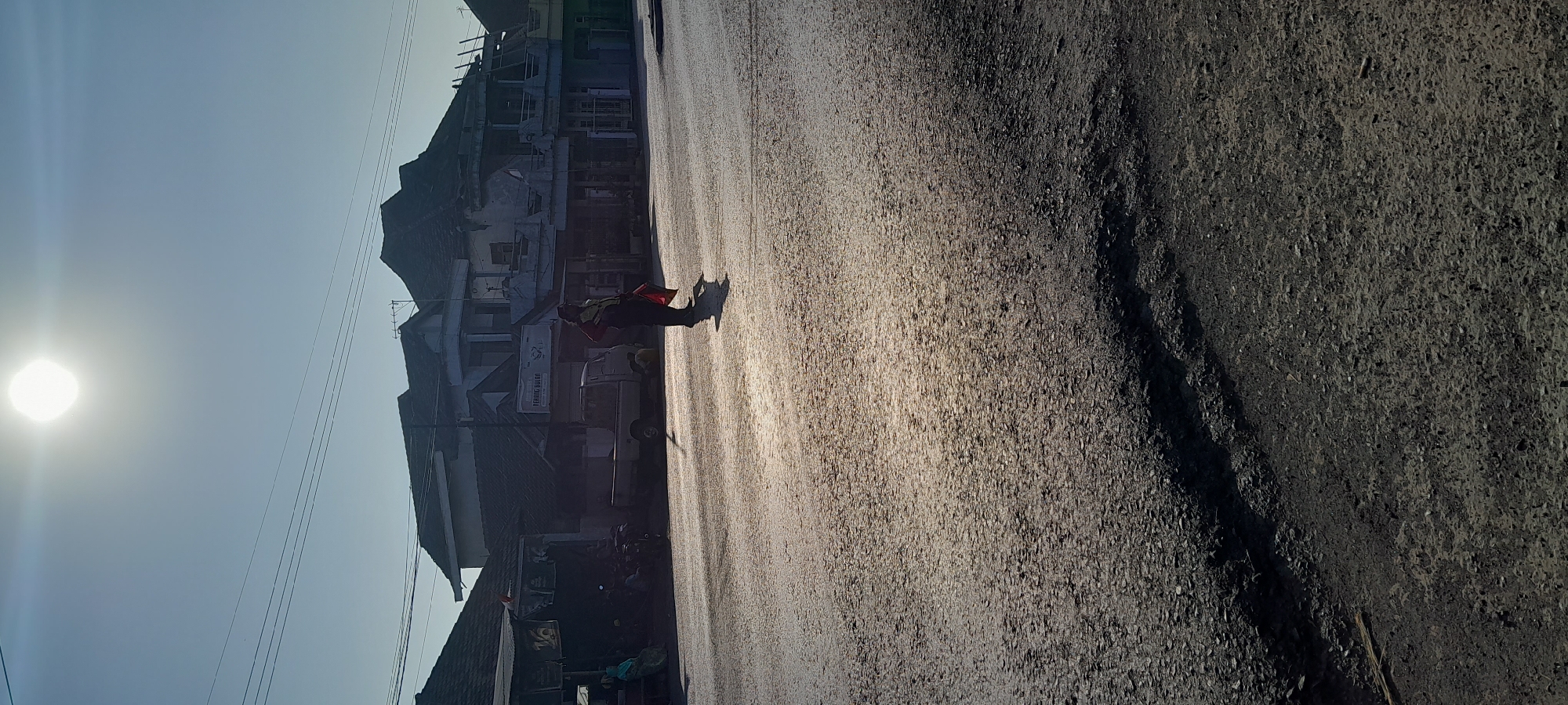 The image depicts a street scene in Tambakrejo, Indonesia, under a bright, hazy sun. The foreground is dominated by a rough asphalt road surface with visible patches of damage. In the mid-ground, a person wearing a hat and a red and yellow vest is walking across the road, holding a red flag. Behind them, a light-colored utility vehicle is parked. To the left of the vehicle, a row of buildings lines the street, appearing to be shops or residences. Some motorcycles are parked in front of these buildings. A woman wearing a light blue headscarf sits on a bench to the far left. The background shows more buildings with dark tiled roofs and power lines stretching across the sky. The overall lighting suggests it is either early morning or late afternoon, with the sun casting long shadows.