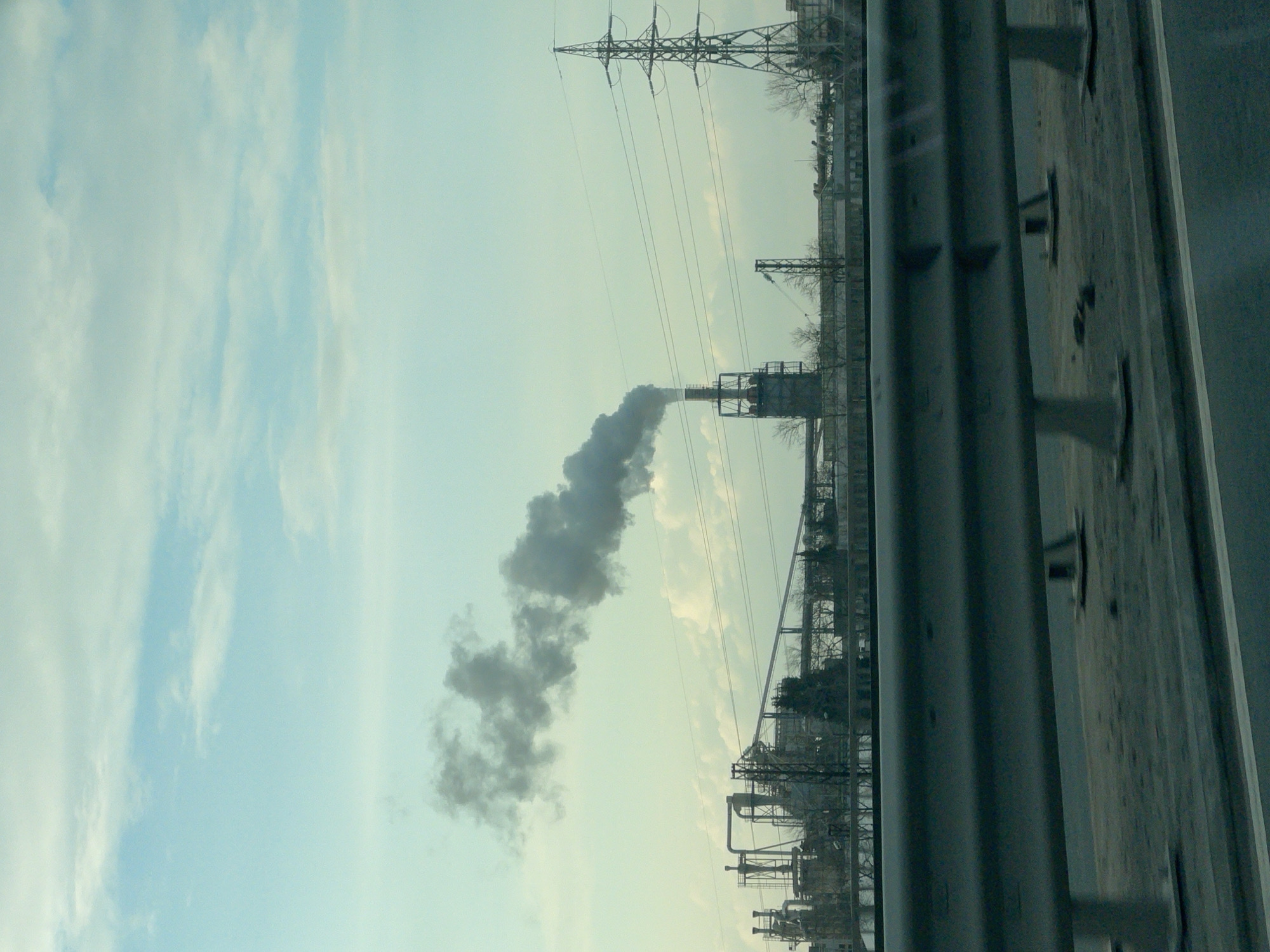The image, likely taken from a moving vehicle, shows an industrial landscape under a pale, overcast sky. Dominating the view is a plume of gray smoke rising from a tall industrial chimney within a factory complex. Power lines stretch across the horizon, supported by a tall pylon. A gray concrete barrier, possibly part of a highway, occupies the right side of the frame. The location is possibly Burgas, Bulgaria, given the style of industrial construction in the area.