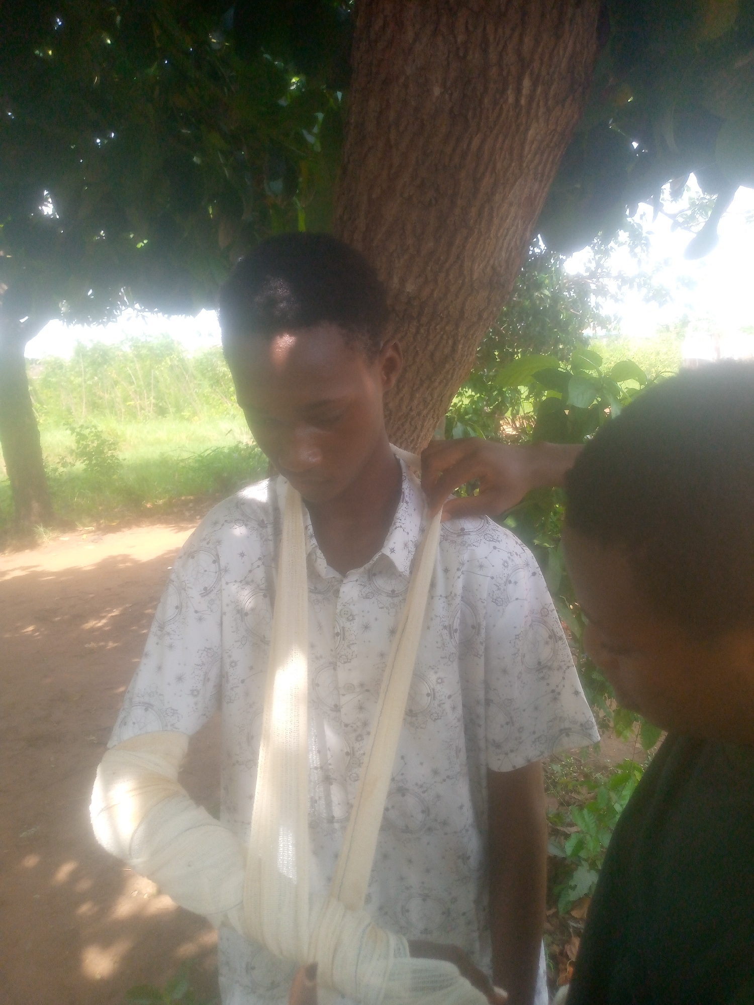 The image shows a young man with a bandaged left arm standing outside. A person is adjusting the sling that supports his arm. The location appears to be a rural area, possibly near a tree or in a park. The city and specific location are unknown.