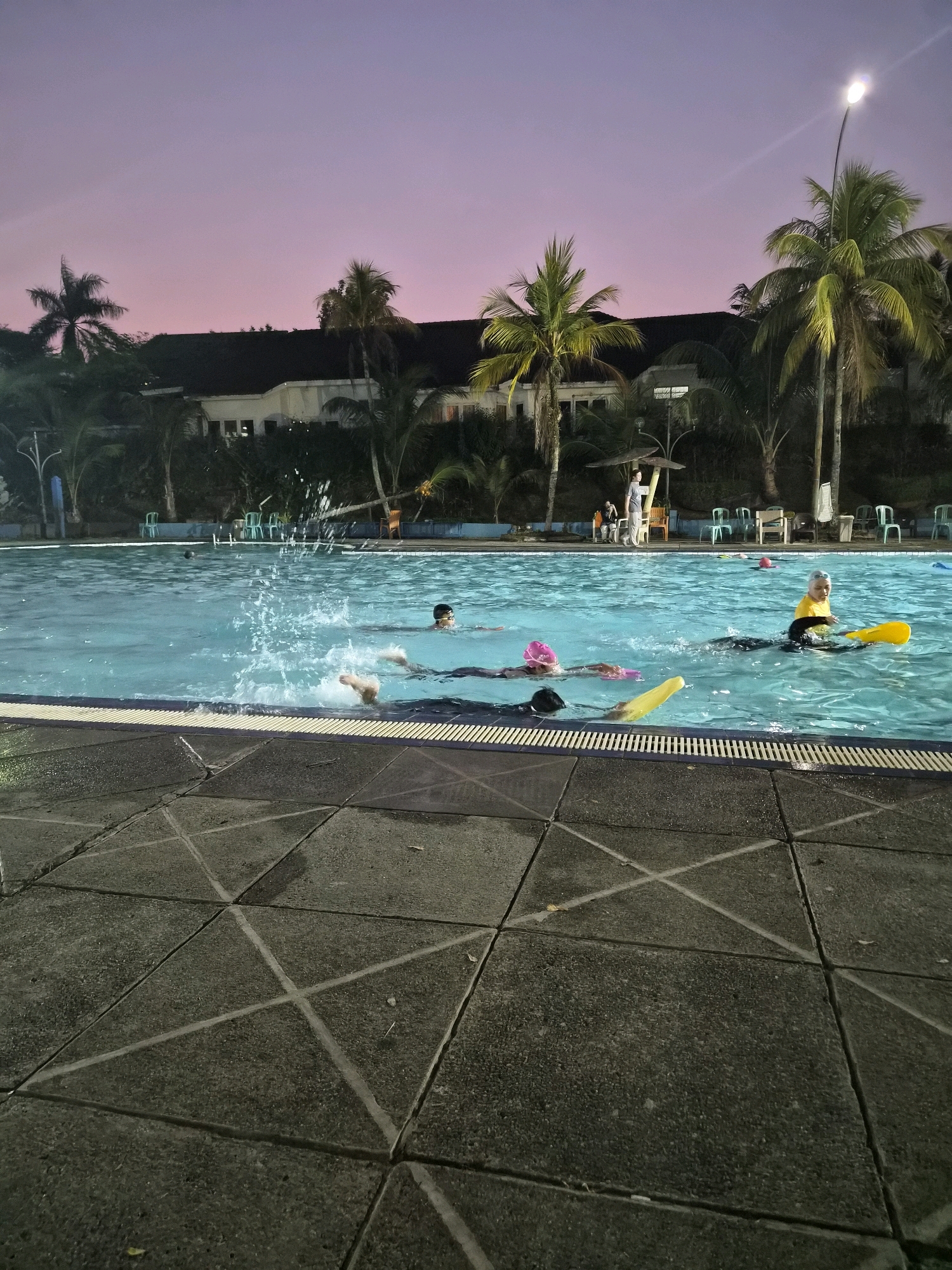 The image depicts an outdoor swimming pool at dusk, likely located in Balikpapan, Indonesia, given the presence of palm trees and the architectural style of the building in the background. The sky shows a gradient of pink and purple hues. Several people are in the pool, engaging in various activities. One individual, wearing a yellow top and black bottoms, is using a yellow kickboard. Another person, with a pink swim cap, is also in the water. There are visible splashes in the water, suggesting movement and activity. The pool is surrounded by a tiled walkway with a geometric pattern. In the background, there is a building with a dark roof, and more palm trees are scattered around the property. A streetlamp is also visible, providing illumination. The overall scene suggests a recreational setting during twilight hours.