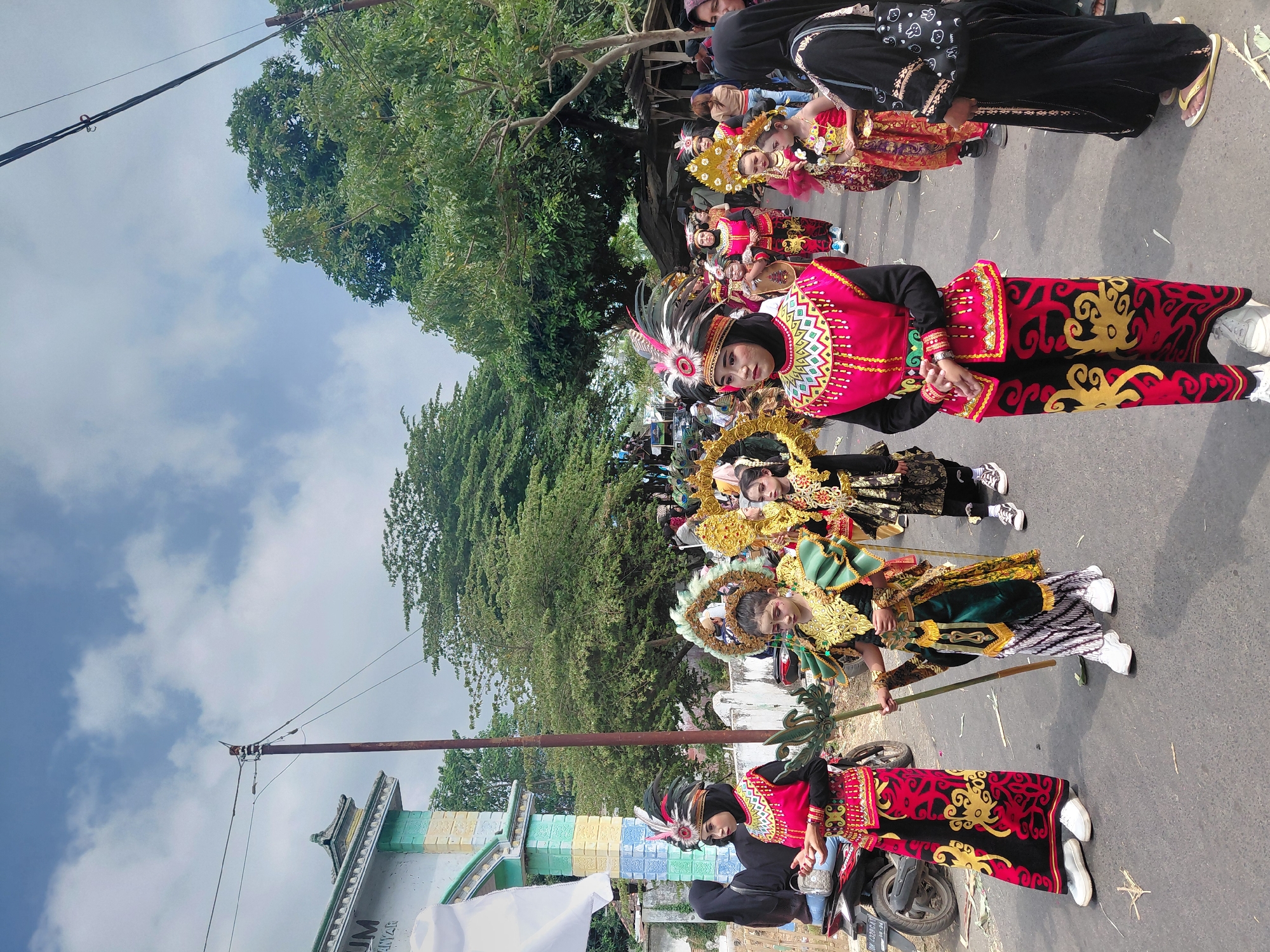 In Bimo, Indonesia, a procession is underway on a paved road under a partly cloudy sky. Several young women and girls are participating, dressed in elaborate, traditional attire featuring vibrant colors and intricate patterns. The costumes include headdresses adorned with feathers and metallic embellishments, ornate tops, and long, patterned skirts or pants. Some participants carry decorative props, such as staffs or fan-like objects. The procession is moving from left to right in the frame. In the background, trees and buildings are visible, including a building with text that appears to say "UM." A utility pole with wires stands on the left side of the road, and a motorcycle is parked nearby. Spectators can be seen observing the event from the sides of the road. The scene captures a cultural event likely a parade or festival.