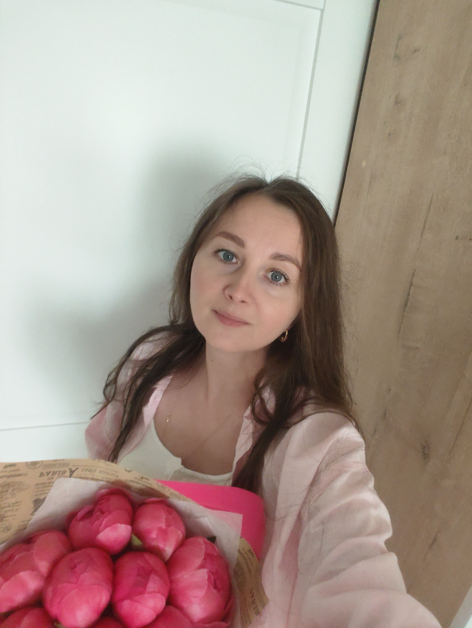 A young adult woman with long, medium-brown hair and light blue eyes is shown in a close-up, slightly high-angle shot, looking directly at the camera with a neutral to subtly pleasant expression. She is wearing a light pink, possibly silk or satin, button-up shirt unbuttoned over a white top, revealing a delicate gold chain with a small pendant. She also wears small gold hoop earrings.
She holds a bouquet of numerous tightly-budded pink flowers, likely peonies or tulips, wrapped in rustic brown paper. The paper features faint, decorative text print, some of which is illegible but includes fragments resembling "CREST SOUVENIR" and other stylized characters. A bright pink material forms the base or a ribbon accent for the bouquet.
The background on the left consists of a plain white wall or door, while the right side shows a light brown wooden panel or surface. The lighting is soft and even, indicating an indoor setting.
