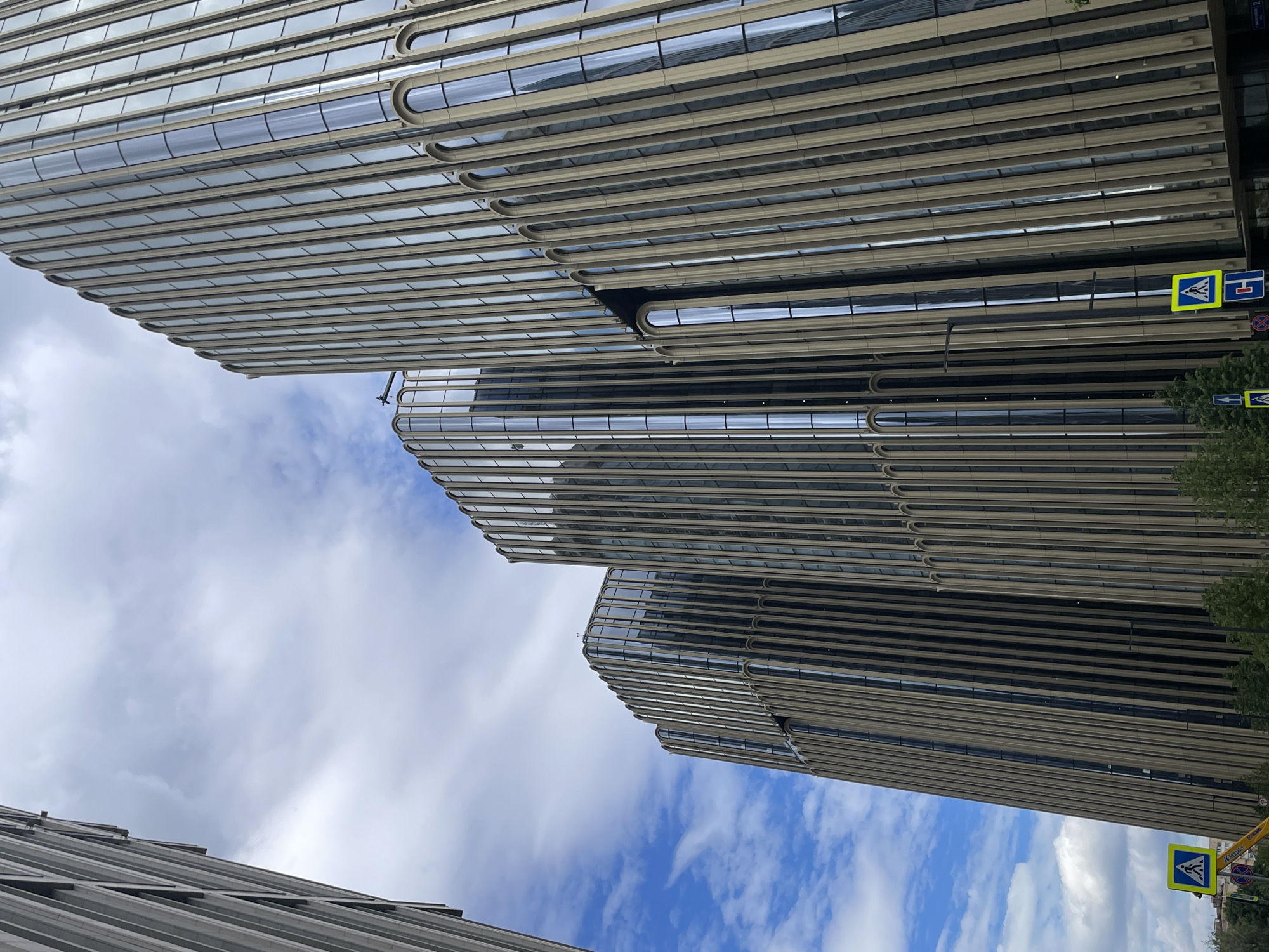 The image shows the contemporary architecture of a row of tall, slender skyscrapers with a unique design featuring vertical golden ribbed facades and arched glass windows. The buildings are situated in a city environment, indicated by the presence of traffic signs, including a pedestrian crossing sign. The sky is partly cloudy with blue patches, suggesting it is daytime. The architecture implies a modern urban setting, likely in a business district. The buildings appear to be constructed with glass and a metallic material, reflecting the sky. The perspective is from a low angle, looking upwards at the imposing structures. The exact location in Moscow, Russia is not explicitly identifiable from the visual cues alone, however, the style of architecture is consistent with recent urban development in affluent areas of the city.