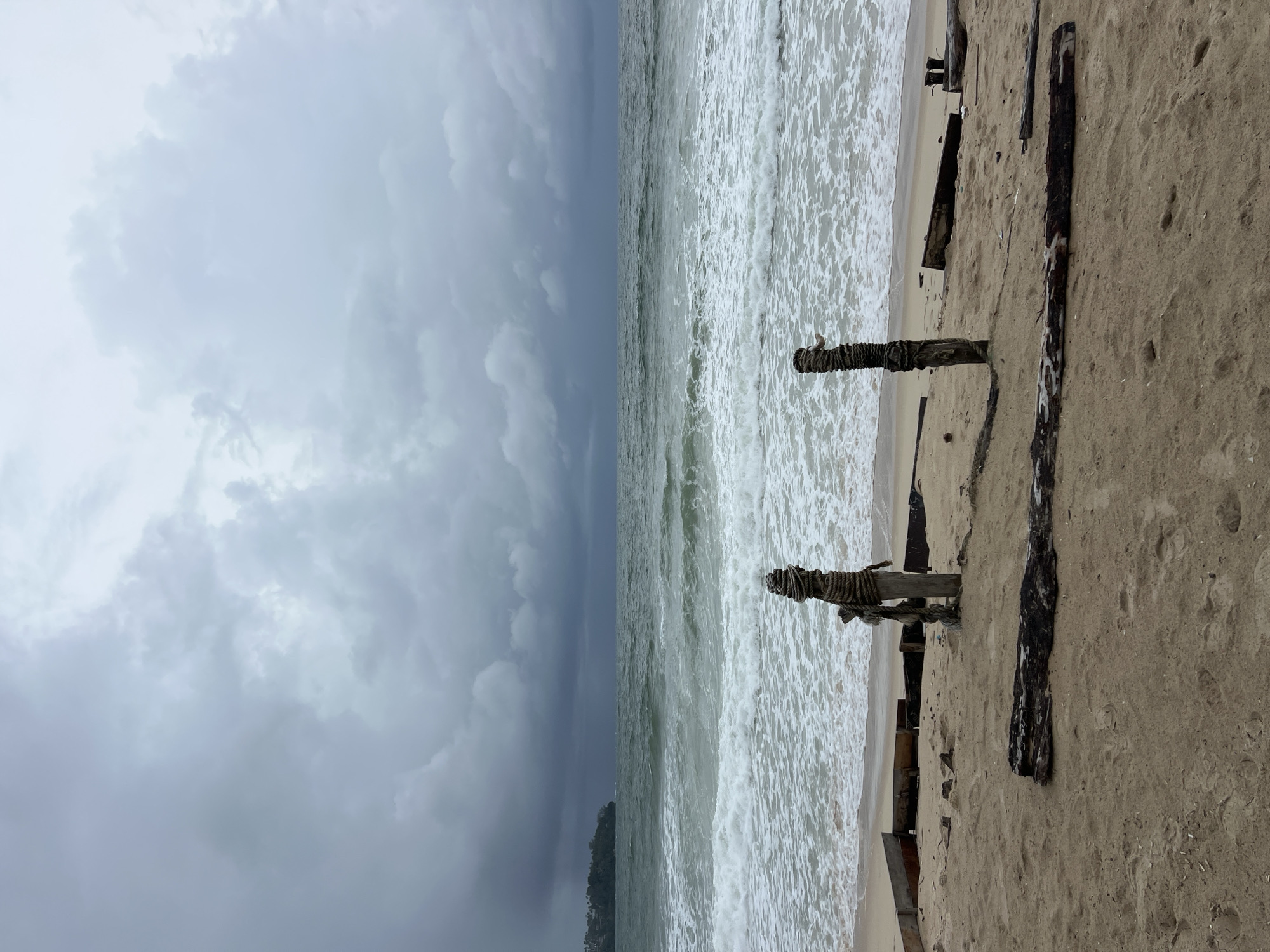 The image depicts a moody coastal scene under a heavy, overcast sky. Dark, voluminous clouds dominate the upper portion of the frame, giving a sense of impending storm or a dramatically cloudy day. The horizon line separates the turbulent, grey-green ocean from the muted sky.
Choppy, foamy waves actively break and crash onto the sandy shore, creating a line of white froth along the waterline. The beach consists of light brown sand, appearing wet near the ocean's edge. Numerous pieces of weathered wood are scattered across the sand. Several dark wooden posts, possibly remnants of an old pier or breakwater, are partially buried in the sand. One prominent post in the foreground has thick, dark ropes tightly wound around its upper section. Long, dark pieces of driftwood or broken timber planks also lie horizontally on the sand, some partially covered by foam from the retreating waves. Faint footprints can be seen in the sand, suggesting human presence earlier, though no people are visible in the scene.
A small, dark landmass, appearing as a distant island or forested hill, is faintly visible on the far horizon, peeking out from beneath the low-hanging clouds. The overall atmosphere is somber and powerful, emphasizing the raw nature of the sea and sky. There is no visible text in the image.