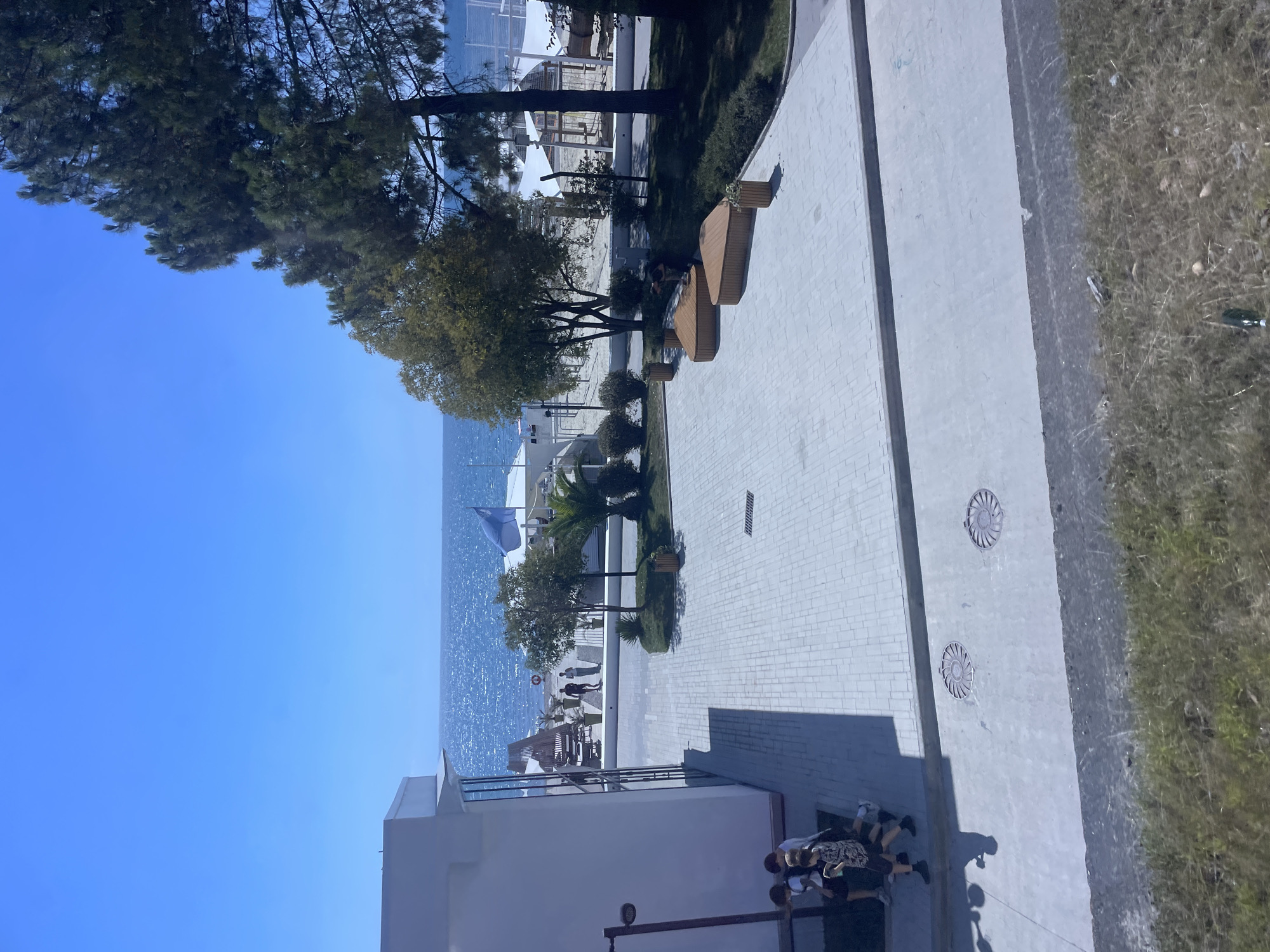 This is a high-angle shot of a modern resort area on the coast of Sochi, Russia, under a clear blue sky. In the foreground, a white building with large glass windows is partially visible on the left. A paved courtyard with a light gray brick pattern leads to the beach area. Several people are present. On the left side of the courtyard, four young males are walking. Further back, two adult males are walking on the paved area near the beach. The beach itself is sandy, with white tents and lounge chairs set up. The Black Sea stretches out to the horizon, shimmering under the sunlight. Several trees, including pine trees, line the edge of the resort area, providing shade. Two triangular wooden benches are placed in the courtyard. A few potted plants are also visible. The overall scene suggests a well-maintained vacation destination.