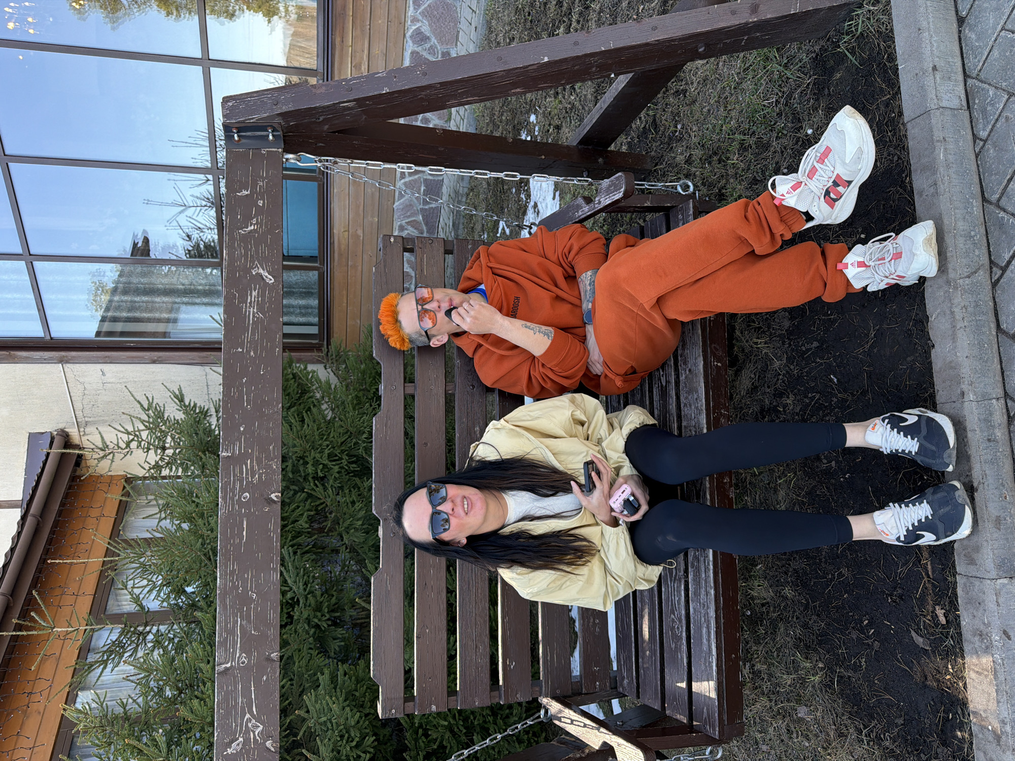 Two individuals, a male and a female, are sitting on a brown wooden swing-bench outdoors. The male, positioned at the top of the frame, has orange hair, is wearing orange clothing, and has tattoos on his arms. He is holding a black object near his mouth. The female, below him, has long dark hair, sunglasses, is wearing a beige jacket over a white shirt with black leggings, and is holding a pink cell phone in her hands. The swing is suspended by metal chains and is situated near a building. The environment consists of a combination of grass, dirt, and paving stones. The location is assumed to be in the city of Votkinsk, Russia, given the prompt's specifications.