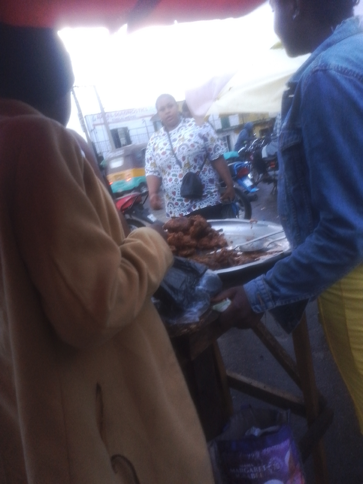 In a bustling street scene in Gwong, Nigeria, two individuals are captured in a candid moment, likely at a market stall. The foreground features a person in a light brown coat, their back mostly to the camera, revealing a glimpse of a shoulder and arm. To their right, another person is visible, wearing a blue denim jacket and yellow trousers. They appear to be interacting with a large metal basin filled with what looks like fried food, possibly chicken or fish, displayed on a wooden table. A serving utensil rests in the basin.
In the background, a woman in a white top with a colorful pattern is standing, holding a dark bag. Behind her, a building with "GLOBAL DIAGNOSTICS" prominently displayed on its facade suggests a commercial area. Several motorcycles and a yellow and blue vehicle are parked nearby, indicating the active transportation and commerce typical of the location. The lighting suggests it is daytime, with bright, diffused light, possibly indicating an overcast sky or open market shade. The overall atmosphere appears lively and informal, typical of a Nigerian street market. A purple bag with visible text, including "MARGARETS", is on the ground near the table.