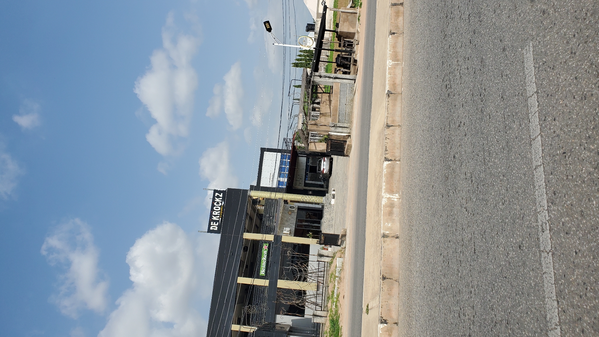 The image captures a sunny daytime scene in Jalingo, Nigeria, featuring the exterior of a two-story building with a dark facade and the prominent signage "DE KROCKZ LUXURY" at the top. The building has several windows and balconies, with some featuring decorative metalwork. A "Heineken" advertisement is visible on the lower level. In front of the building, a paved area with a fence and some outdoor seating structures is present. A white car is parked near the entrance of the building. To the right of the building, a lamppost with LED lights stands beside a street. The foreground shows a paved road with a concrete curb. The sky above is blue with scattered white clouds.