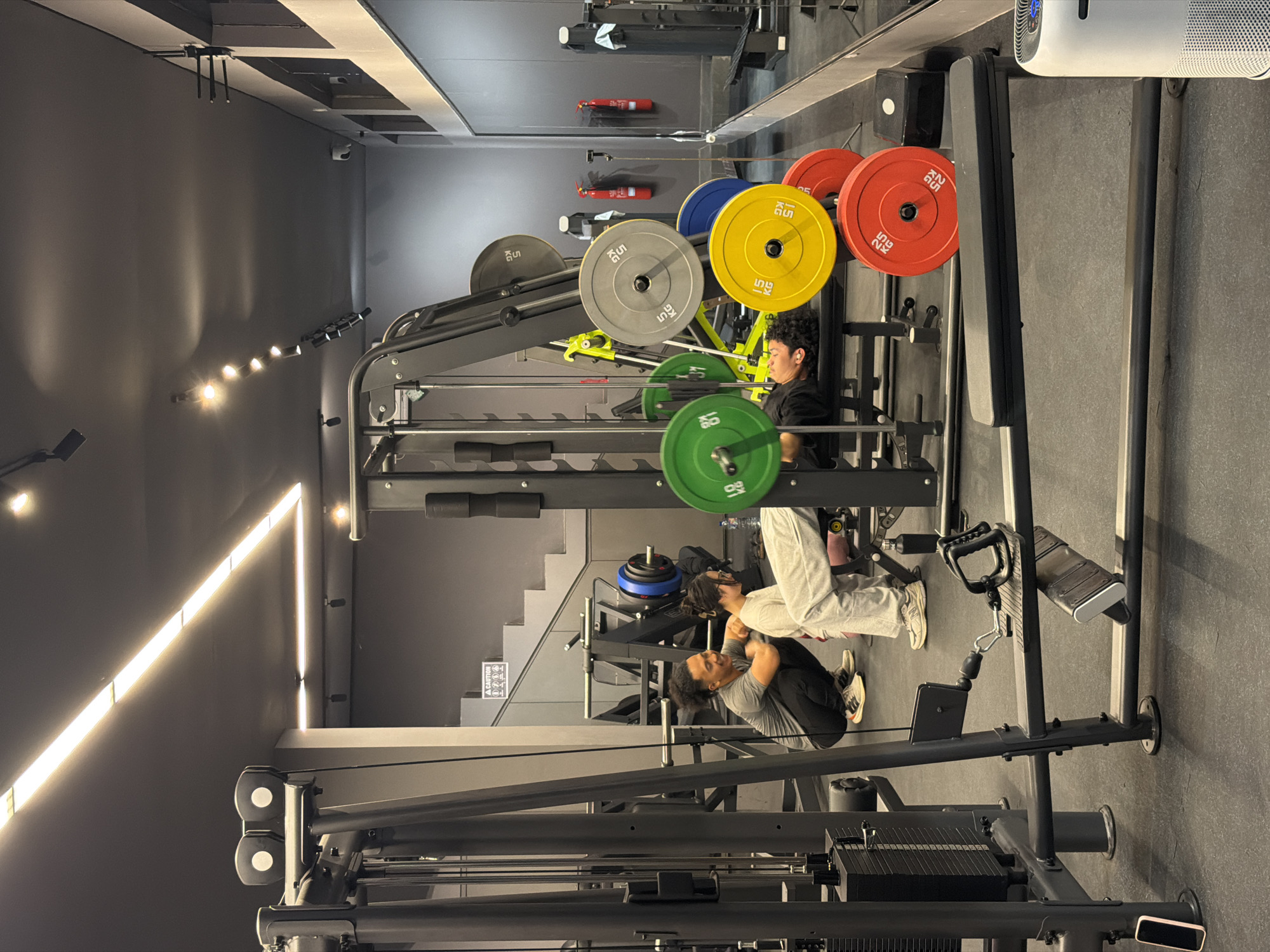 A gym interior is depicted with dark gray walls and ceiling, illuminated by bright fluorescent lights. In the foreground, a black weight bench is prominent. A person lies on the bench, performing a bench press, with colorful weight plates on the barbell. To the left, a man is squatting and looking at the person on the bench, appearing to be offering guidance or encouragement. A child is positioned between them, also watching. Various weightlifting equipment, including a cable machine, weight racks, and barbells with plates, are visible throughout the space. A large mirror lines the right wall, reflecting the gym's equipment. Fire extinguishers are mounted on the wall. The flooring is black rubber.