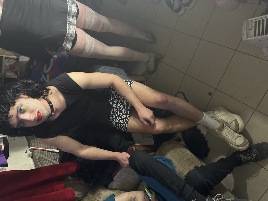 The scene depicts an indoor setting, identified as a private residence in Saratov, Russia. The room features light-colored tiled flooring and is illuminated by dim, artificial light.
A young adult individual is seated centrally, positioned directly facing the camera. This person wears a black wig, heavy makeup including red lipstick and distinct green and blue eyeshadow, a black sleeveless top, and black and white leopard print shorts. A black studded choker is worn around their neck. Their feet are clad in white socks and light-colored sneakers. The individual's left arm extends towards the left, while their right arm rests on their lap, and their legs are crossed at the ankles.
To the immediate right, partially visible from the mid-thigh down, another individual stands. This person wears a short black garment with white lace trim at the hem, sheer light-colored thigh-high stockings, and dark high-heeled shoes.
To the left, a third individual is seated on a light-colored upholstered piece of furniture, with only their lower body visible. This person wears dark athletic pants, dark athletic shoes, and a blue top, with a partial tattoo visible on their arm.
In the background, a dark red fabric hangs vertically, and a white object, possibly a radiator or wall fixture, is present, from which a candy cane ornament hangs. The floor in the foreground shows another pair of sneakers and a white ventilated object. The overall environment appears informal and somewhat cluttered.