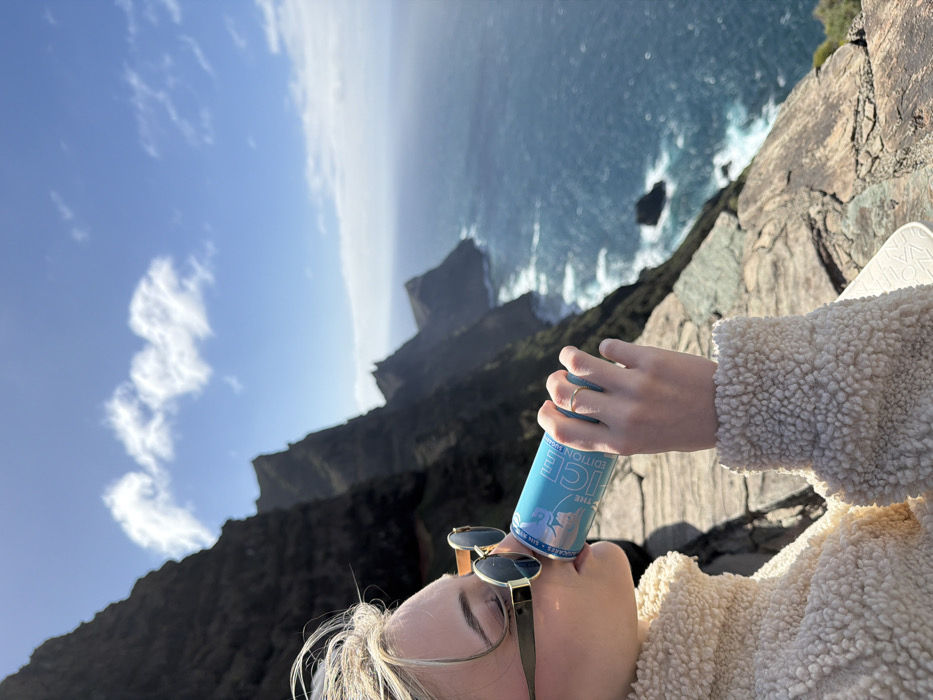 A close-up, side-profile view captures an individual drinking from a light blue aluminum beverage can on a cliffside overlooking the ocean in Tirma, Spain. The person, with blonde hair partially visible, wears dark, round-framed sunglasses and a cream-colored, textured sherpa-style jacket. Their left hand, displaying a thin gold-colored ring on the ring finger, grips the can which bears the text "THE ICE EDITION" and a polar bear graphic. The background features steep, dark rocky cliffs receding into the distance along a coastline. The deep blue ocean, marked by white-capped waves, stretches towards the horizon under a bright blue sky with scattered white clouds. The immediate foreground consists of flat, natural rock formations where the person is positioned.
