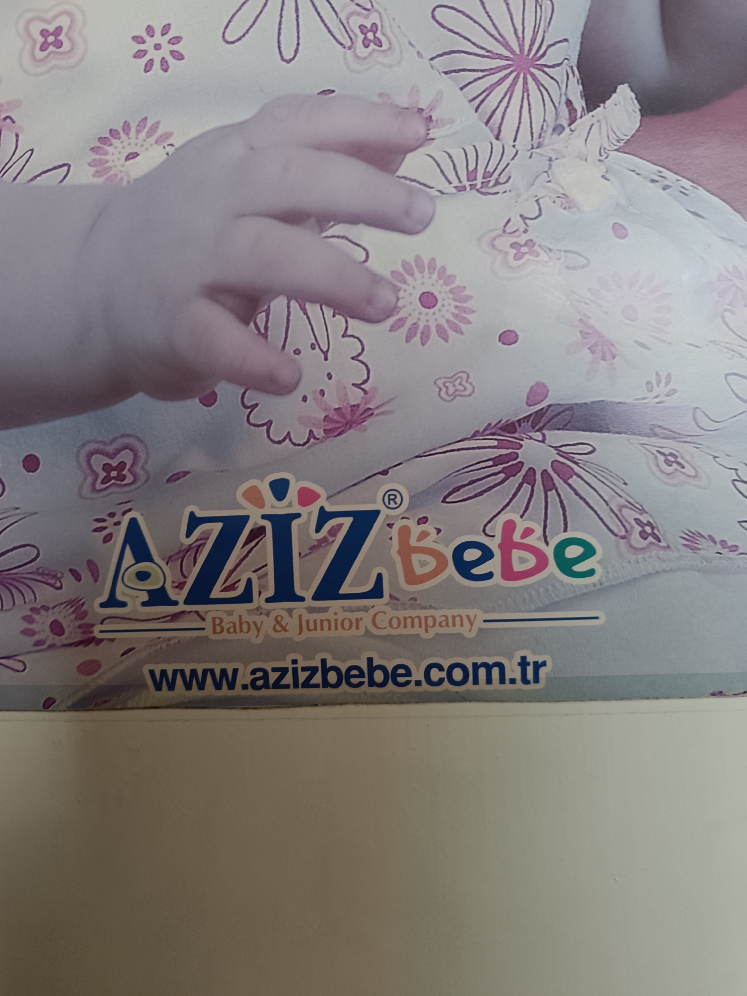 The image shows a close-up of a baby's hand resting on a piece of fabric with a floral pattern. The logo for "AZIZ Bebe Baby & Junior Company" is prominently displayed, along with the website address "www.azizbebe.com.tr". The background appears to be a light-colored surface. There is no information to determine the location in Pleven, Bulgaria, or any other specific location.